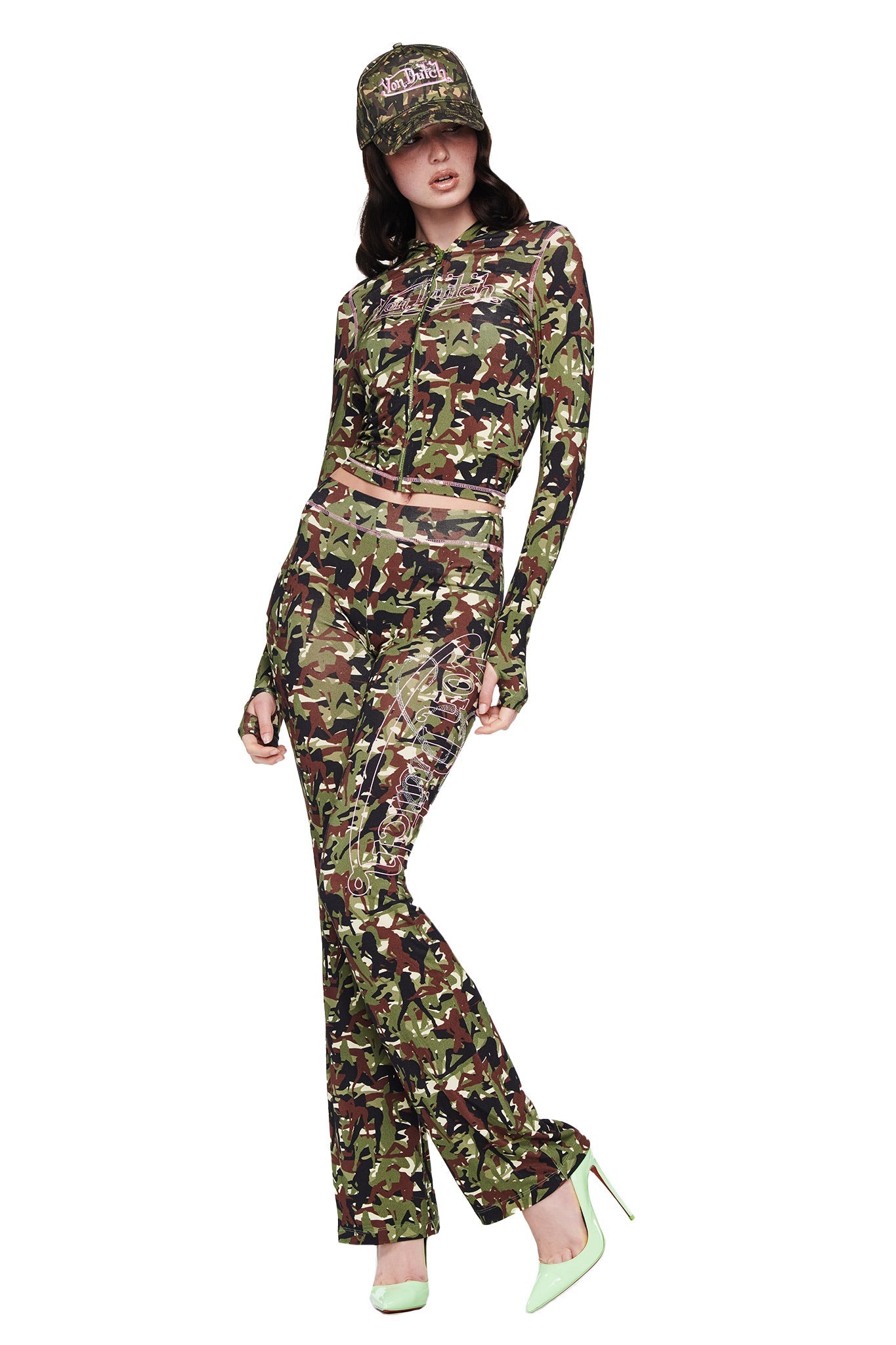 A woman poses in I.AM.GIA's I.AM.GIA X VON DUTCH BLARE TRACKPANT - GIA GIRL CAMO with a matching crop top, Von Dutch cap, and light green heels. The fitted long sleeves and flared pants pop against the white background.