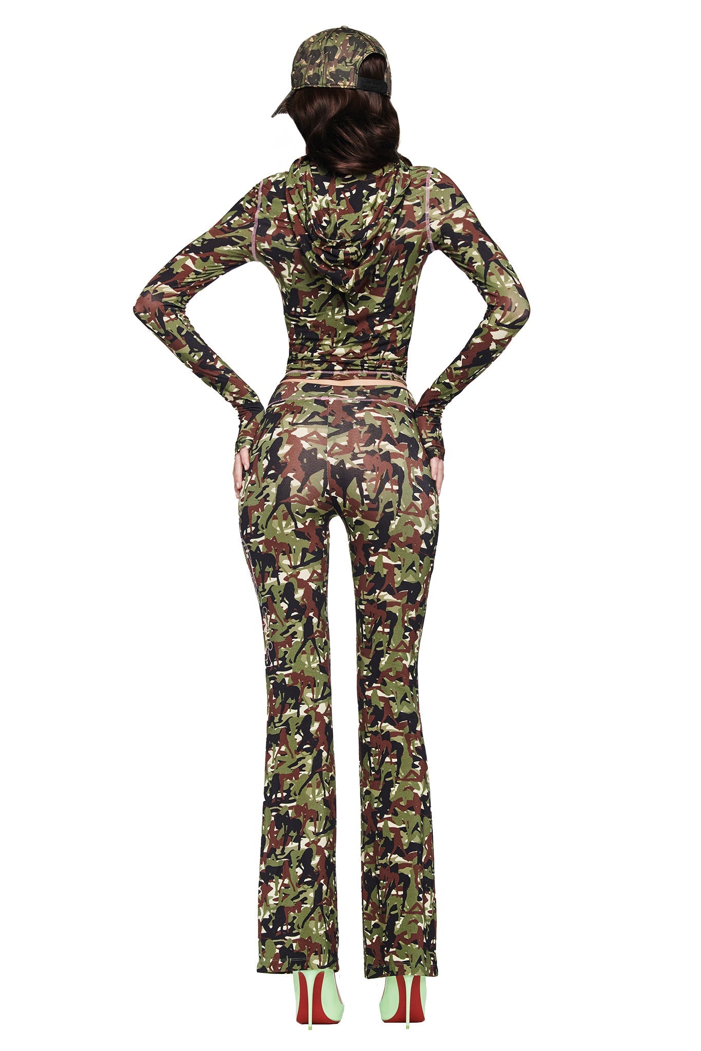 A person with long dark hair in a low ponytail wears the I.AM.GIA X VON DUTCH BLARE TRACKPANT - STRIPPER CAMO set, paired with a matching hoodie, cap, and high heels with green soles, standing against a white background.