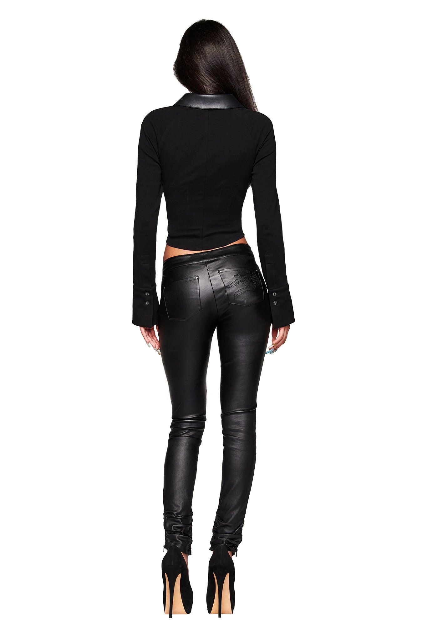 A woman with long dark hair, seen from behind, wears the I.AM.GIA JETT PANT in black with a fitted jacket and heels against a white background.