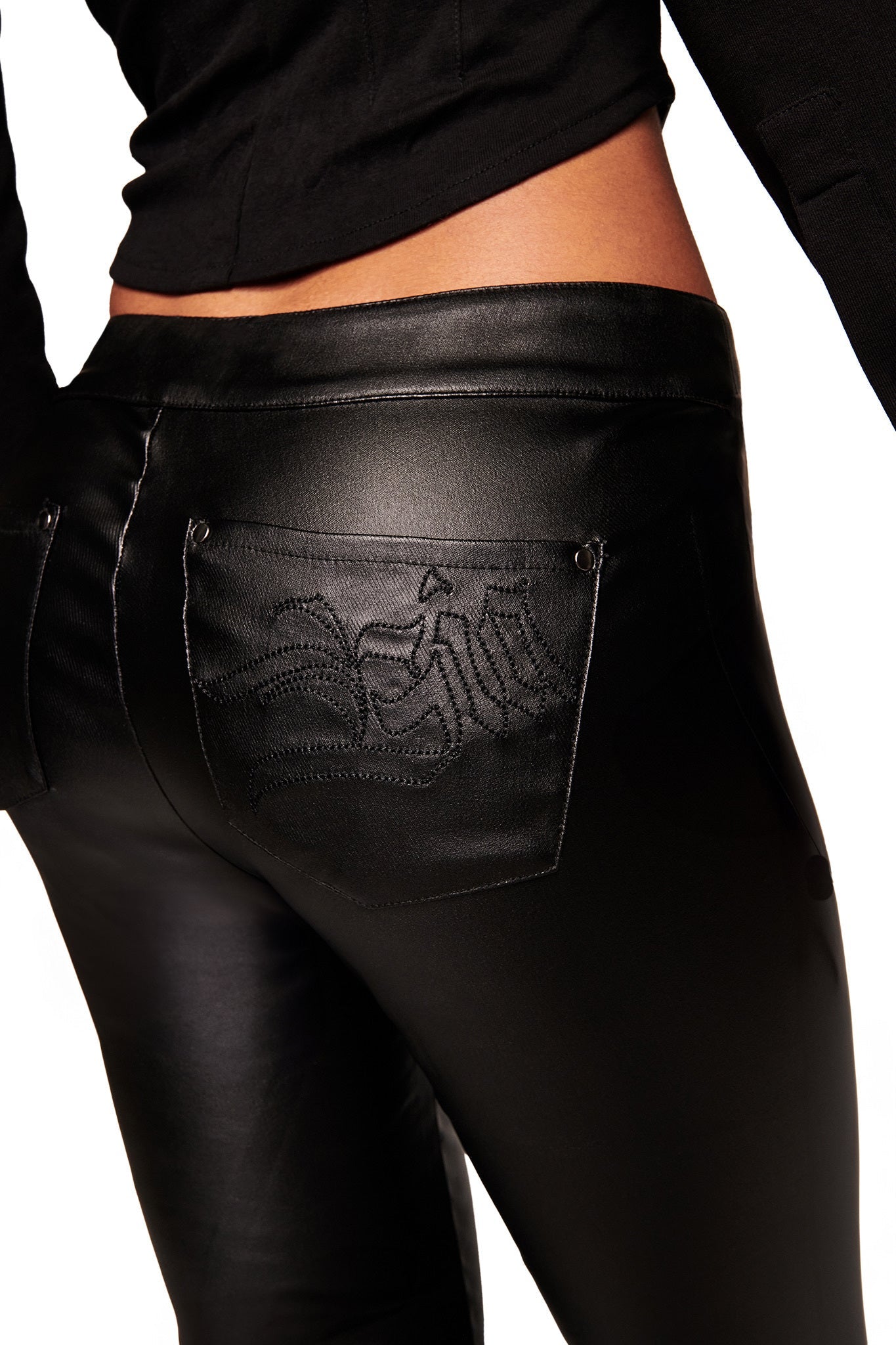 A close-up of a person wearing the I.AM.GIA JETT PANT - BLACK, featuring decorative back pocket stitching. The image highlights the lower back and upper legs, paired with a black top.