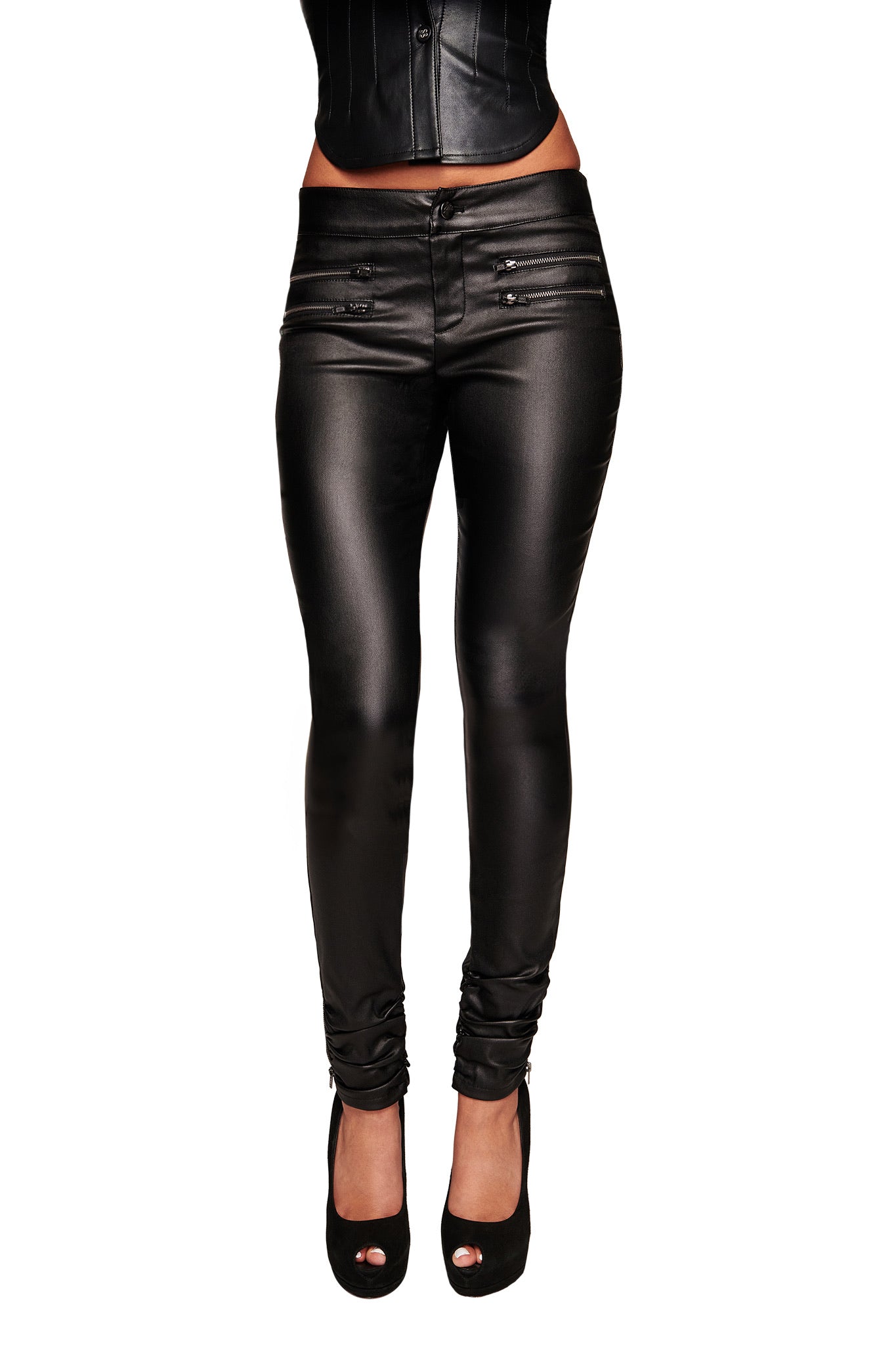 A person modeling the I.AM.GIA JETT PANT - BLACK with a sleek waxed finish, paired with a black corset-style top and black peep-toe heels, shown from the shoulders down against a white background.
