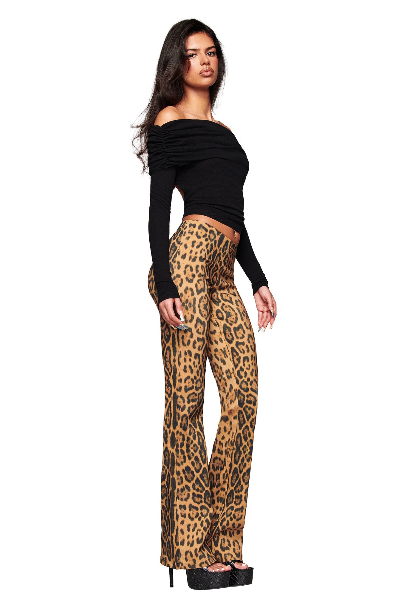 A woman stands in profile against a plain white background, wearing I.AM.GIA’s UMA PANT - PANTHERA animal print flared pants, a black off-the-shoulder top, and black platform heels. She has long dark hair and a confident expression.