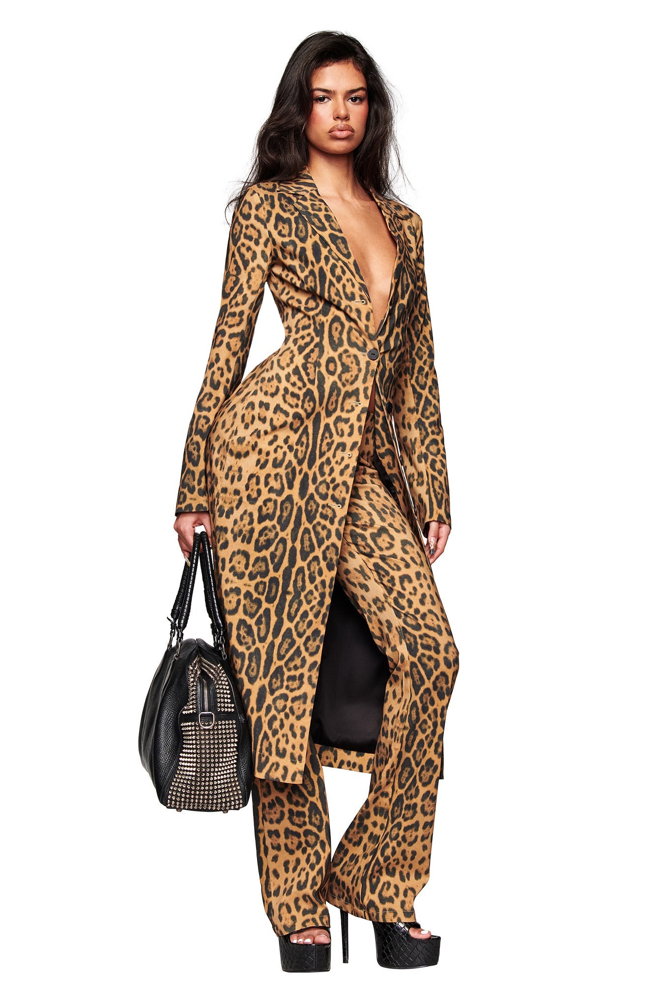 A woman showcases the I.AM.GIA UMA PANT - PANTHERA, featuring a bold animal print and flared legs. She pairs them with a black handbag and platform heels, standing confidently against a crisp white background.