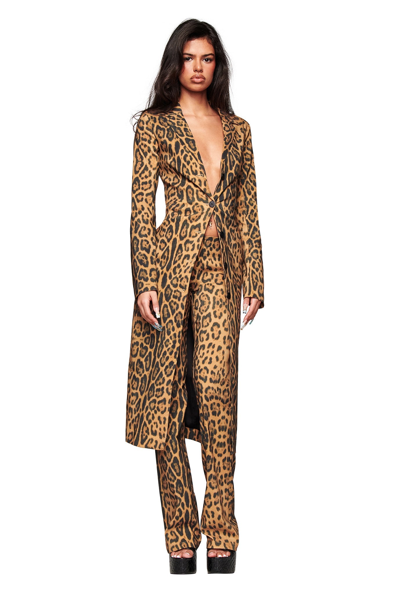 A woman stands against a white background wearing the I.AM.GIA UMA PANT - PANTHERA animal print suit with a long, deep V-neck blazer, flared leg pants, and platform shoes.