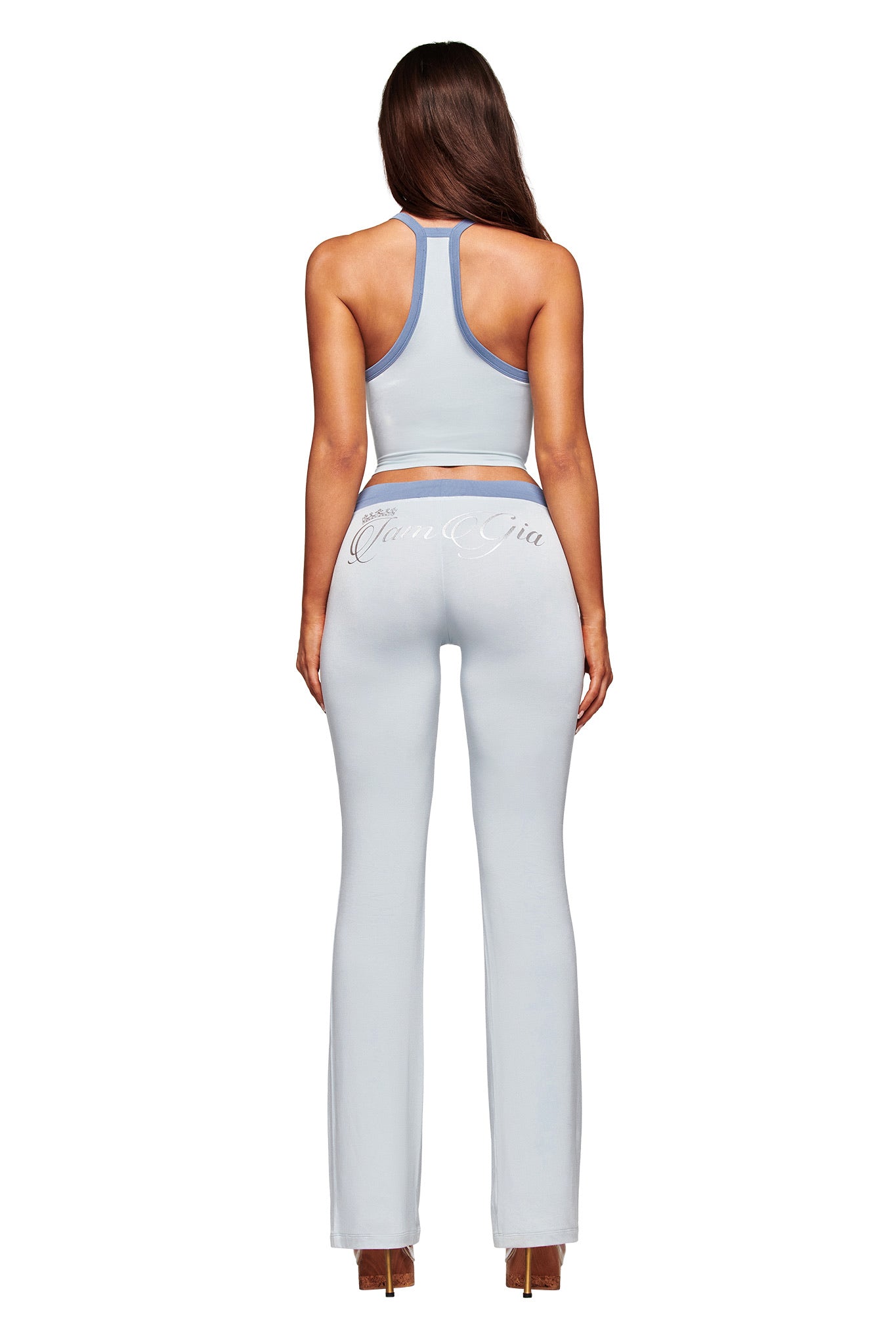 A woman with long brown hair faces away, wearing a light blue sleeveless crop top and I.AM.GIA BLARE LOWER RISE TRACKPANT - TWO TONE POWDER BLUE, paired with high-heeled shoes.