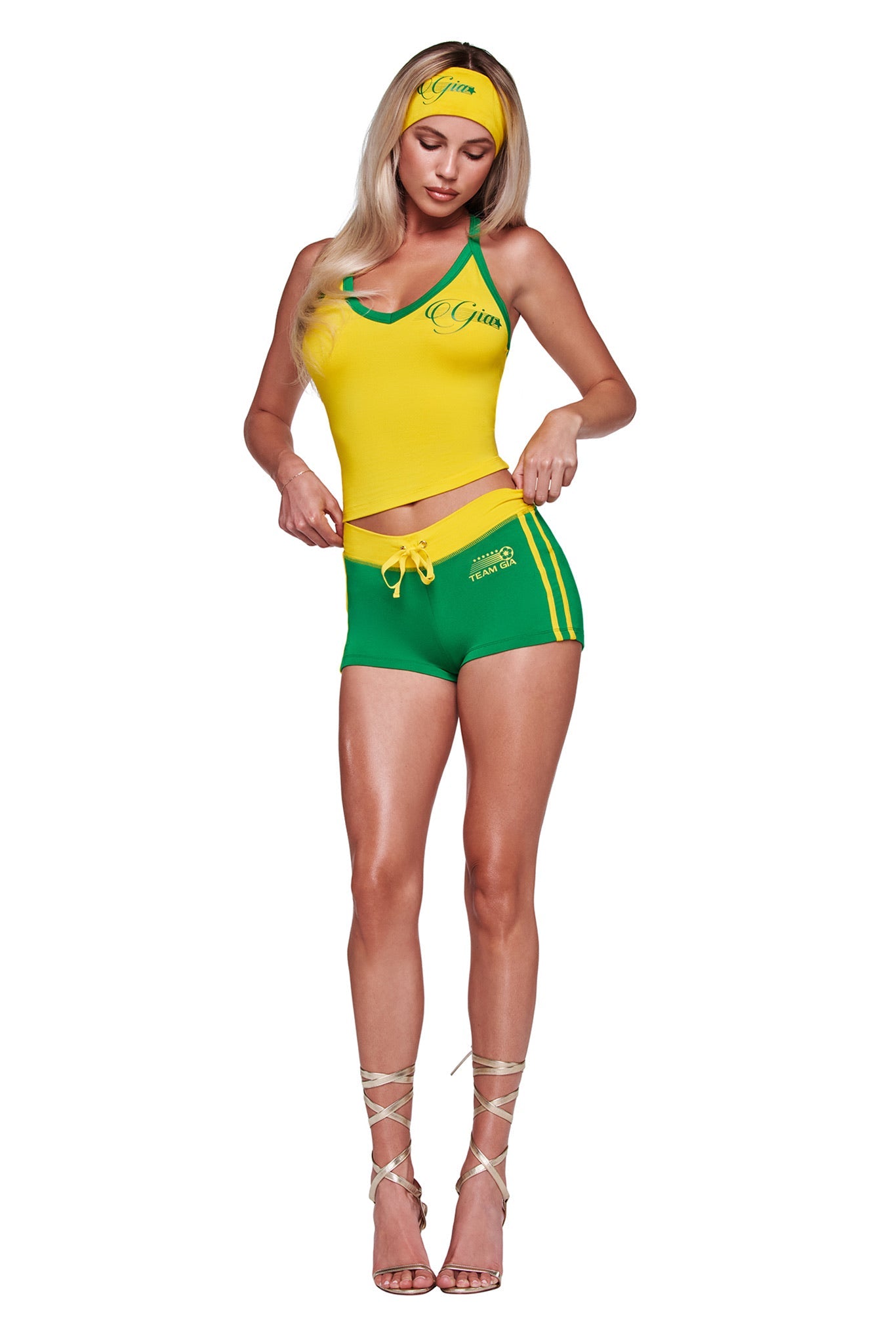 A woman stands against a white background, wearing the I.AM.GIA BLARE V-NECK TOP AND HEADBAND in yellow, paired with matching athletic shorts and strappy high-heeled sandals.