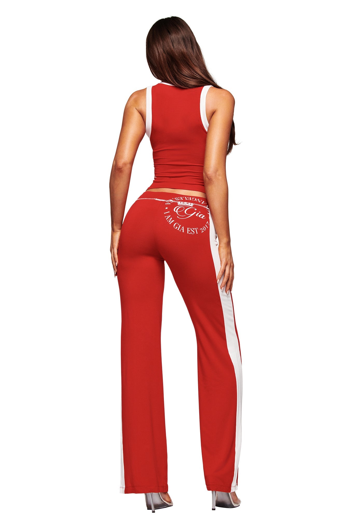 A woman with long brown hair wears the I.AM.GIA BLARE BIND TANK in red, paired with high-waisted bamboo pants and heels, posing with her back to the camera against a white backdrop.