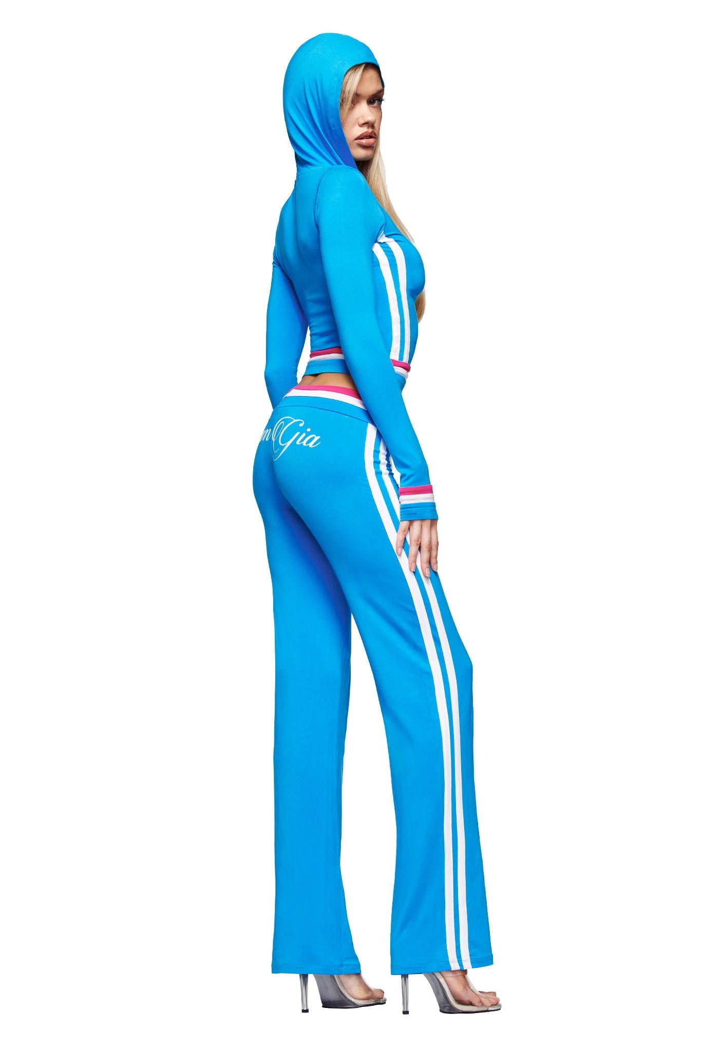 A woman models the I.AM.GIA BLARE STRIPED HOODIE - SKY BLUE, featuring white stripes and a colorful waistband, posing sideways in silver heels against a plain white background.