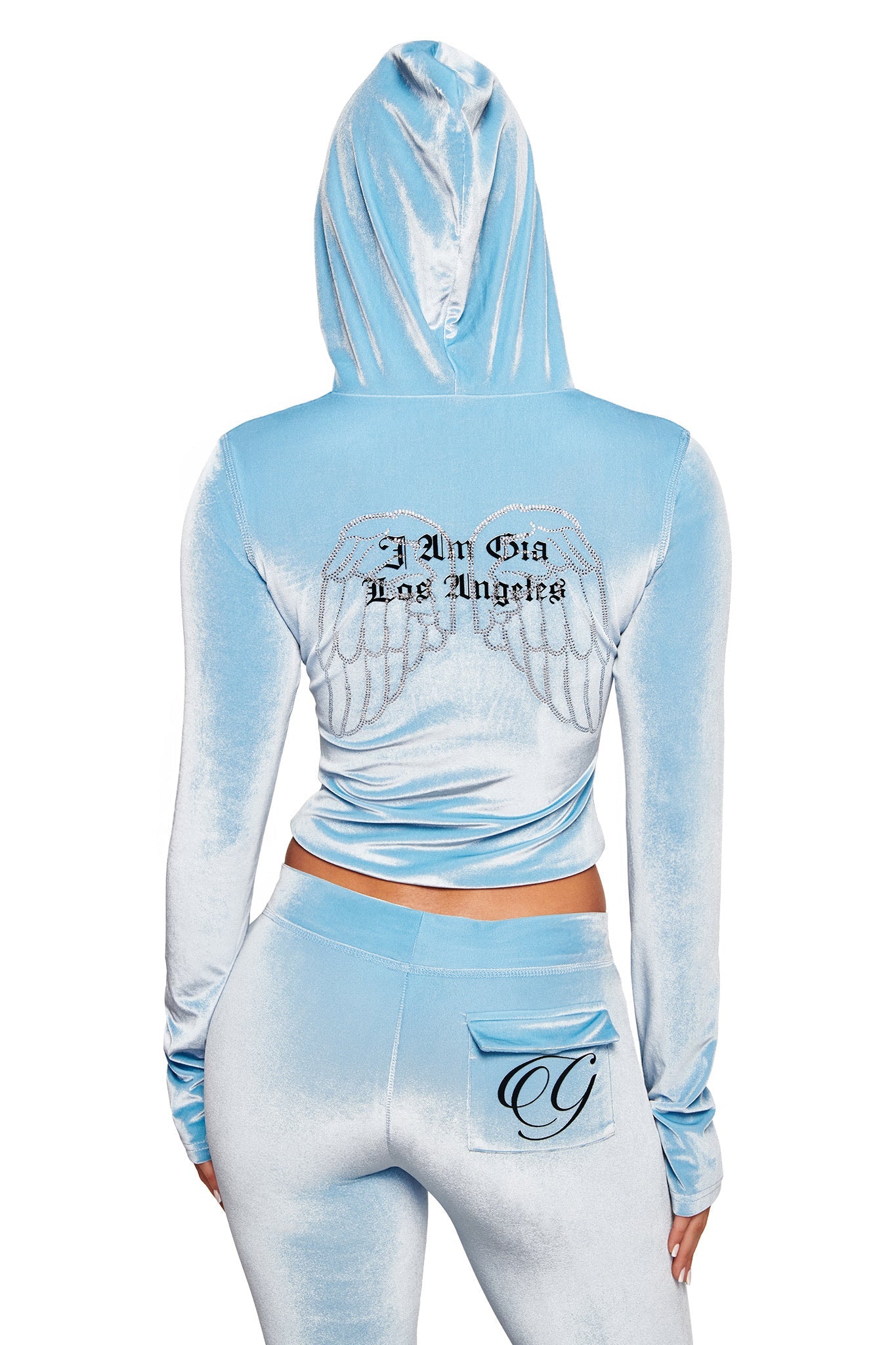 Someone wears the I.AM.GIA BLARE VELOUR POCKET HOODIE in ice blue, featuring diamanté angel wings and “I Am Gia Los Angeles” on the back, paired with matching pants with a stylized “G” pocket detail.