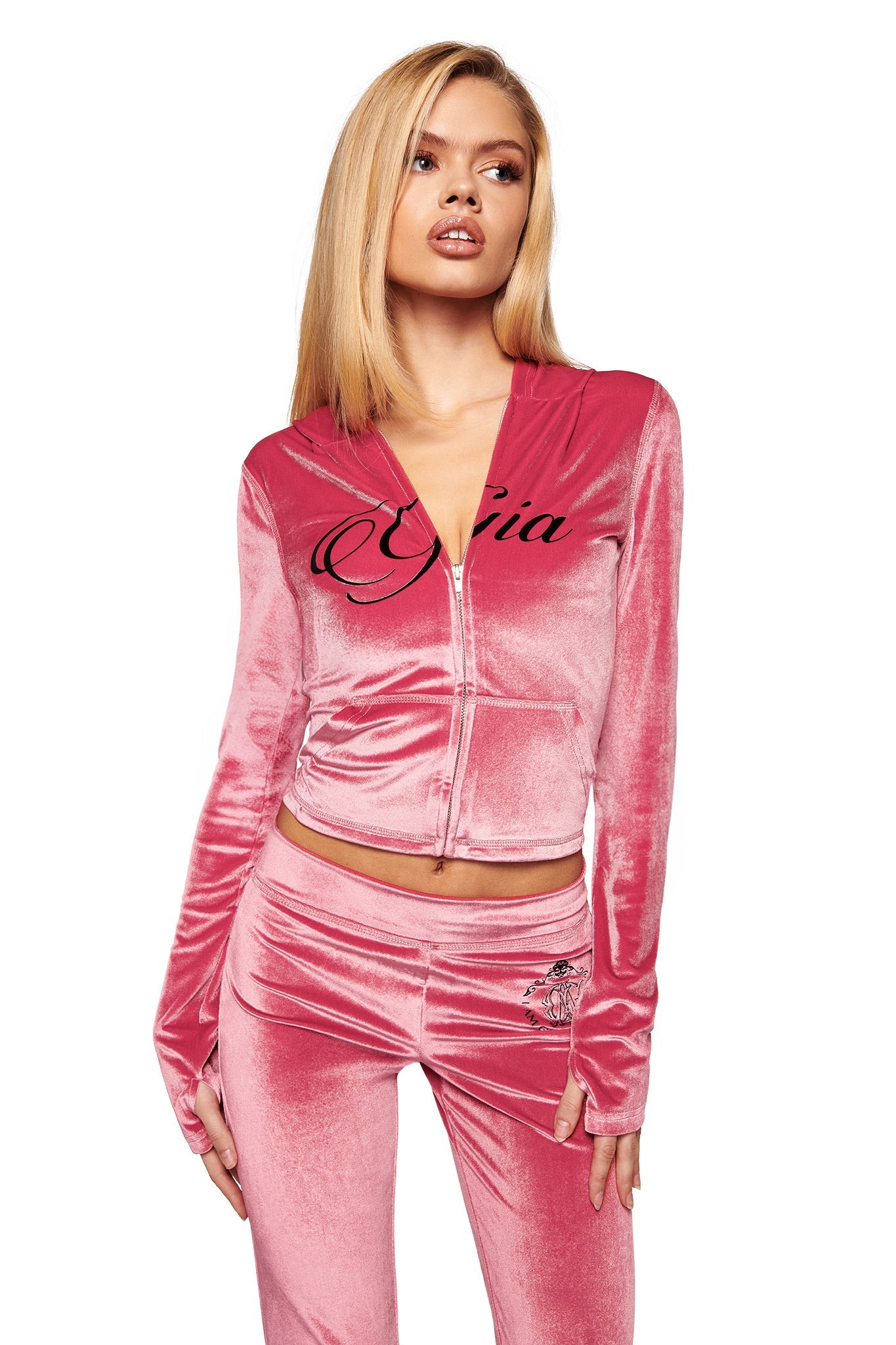 A woman with long blonde hair wears I.AM.GIA's BLARE VELOUR POCKET TRACKPANT in pink, featuring a graphic on the left hip, and a matching zip-front jacket with "Gia" script. She poses against a white background.