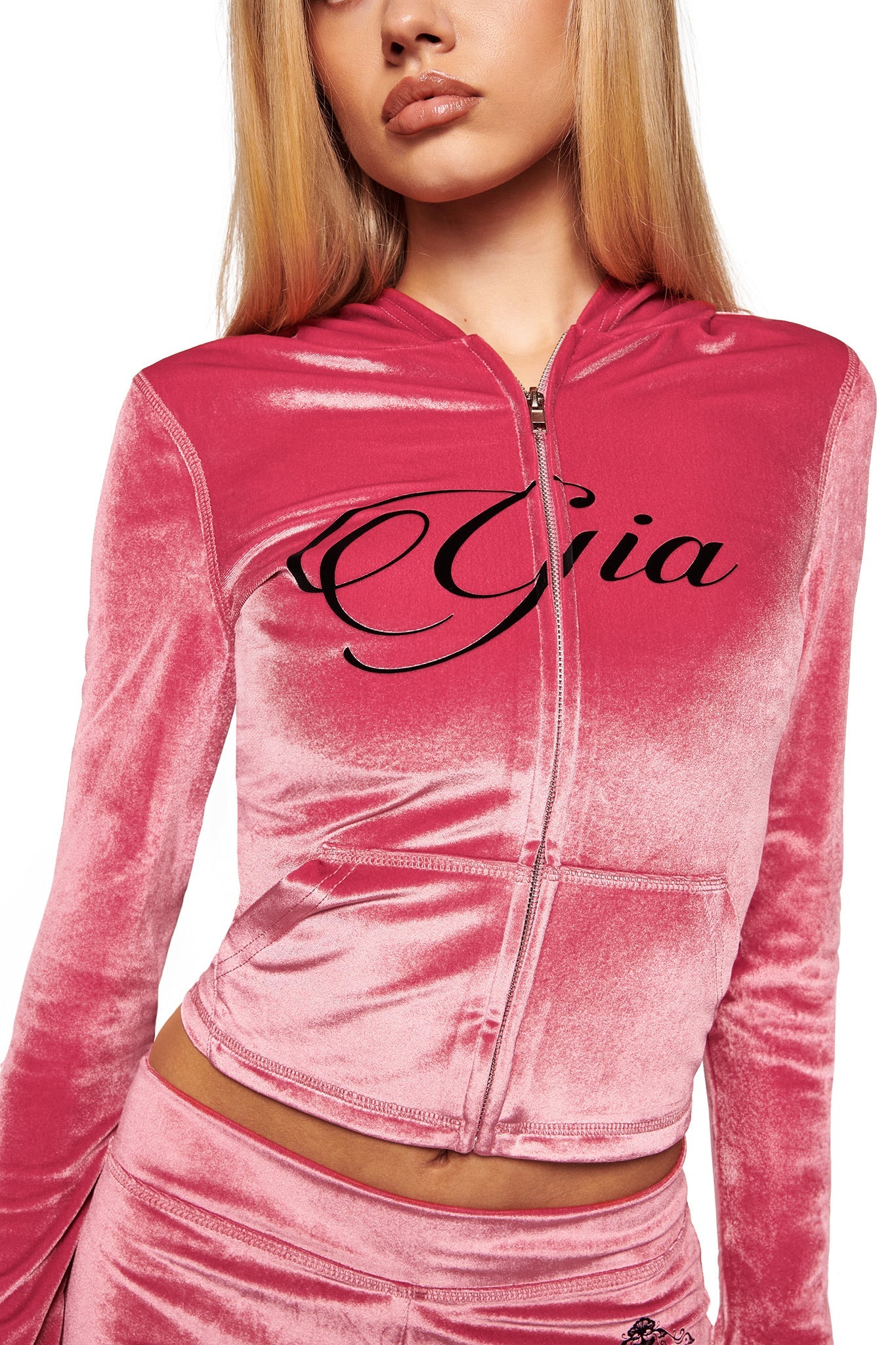 A young woman with long blonde hair wears the I.AM.GIA BLARE VELOUR POCKET HOODIE in pink, with "Gia" in bold black script on the chest and sparkling diamanté angel wings on the back. Only her lower face is shown.