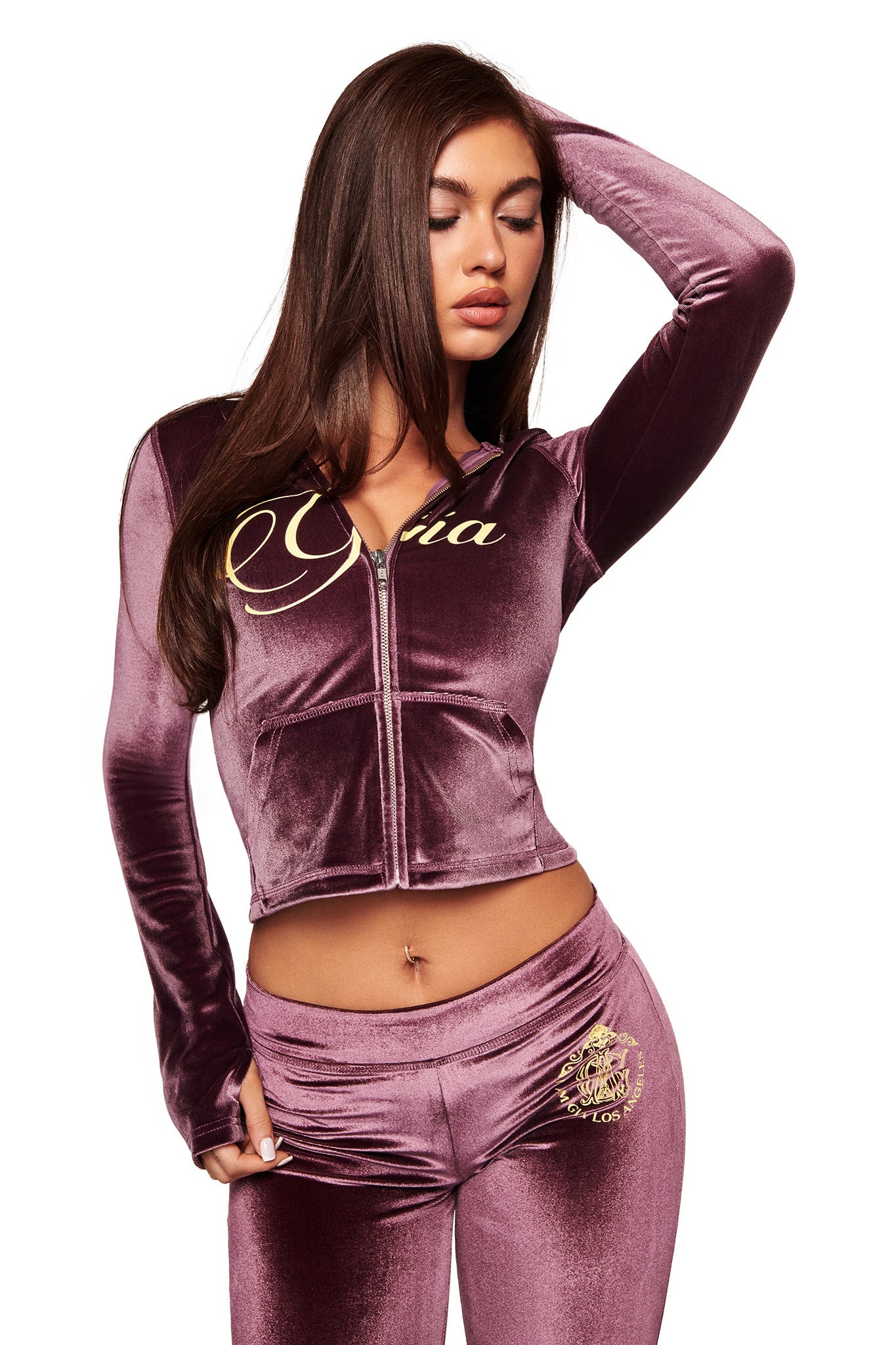 A woman poses against a white background wearing the I.AM.GIA BLARE VELOUR POCKET TRACKPANT in purple, featuring gold GIA script logo.