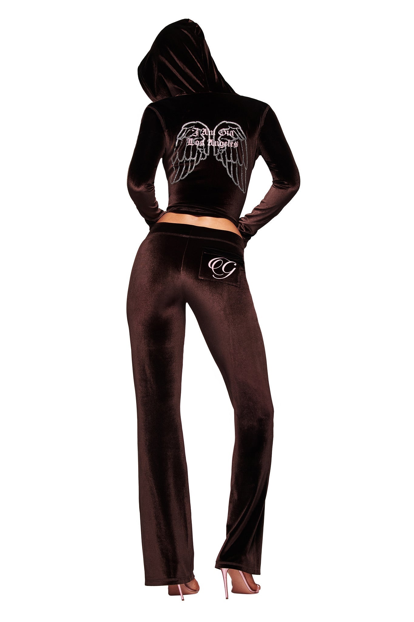 A person wearing I.AM.GIA’s BLARE VELOUR POCKET TRACKPANT in chocolate, featuring a hood and embroidered logo on the pocket, stands in pink heels facing away from the camera.