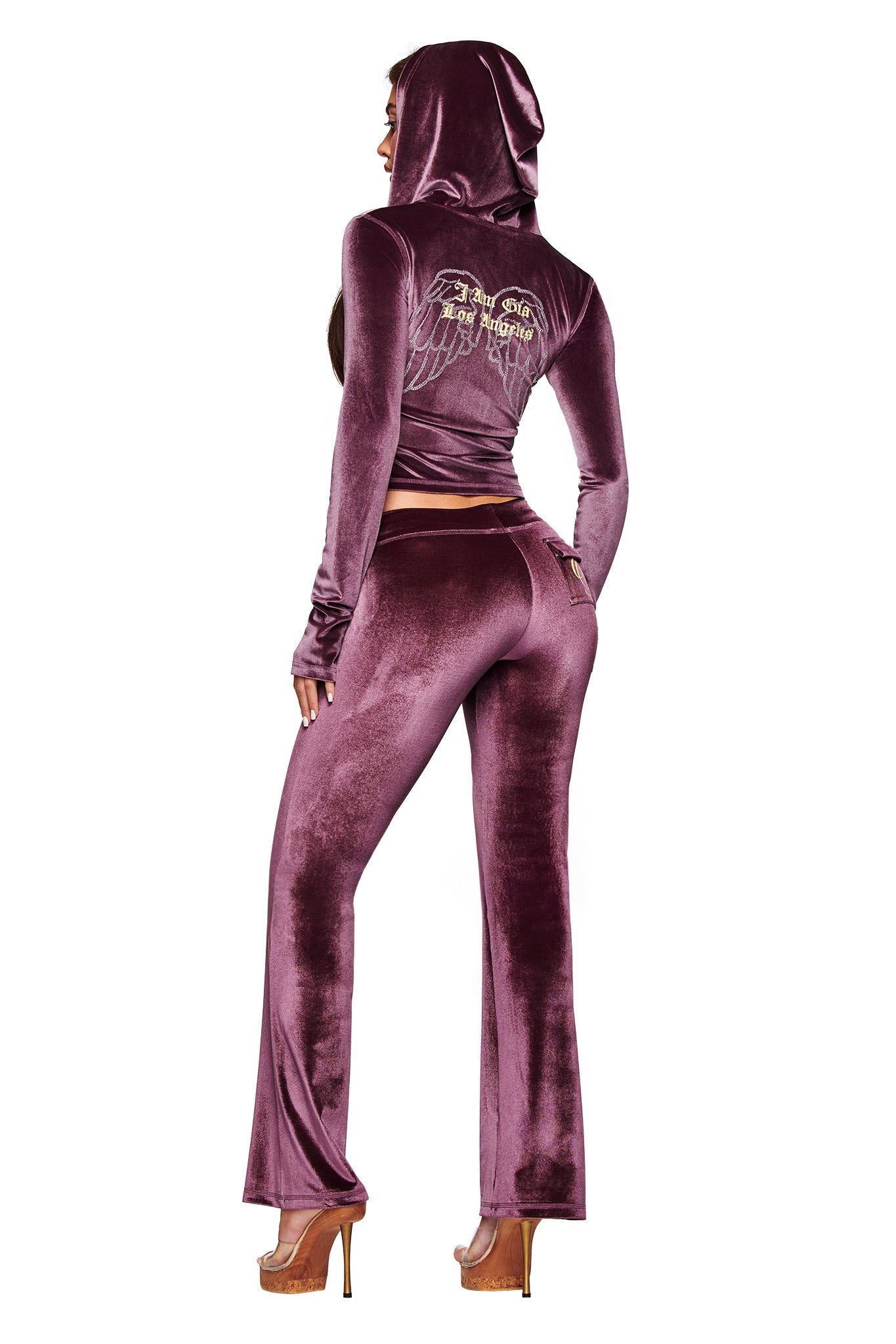 A woman faces away in the I.AM.GIA BLARE VELOUR POCKET HOODIE in purple, featuring diamanté angel wings and the GIA script logo on the back. She wears high platform heels with her hood up.