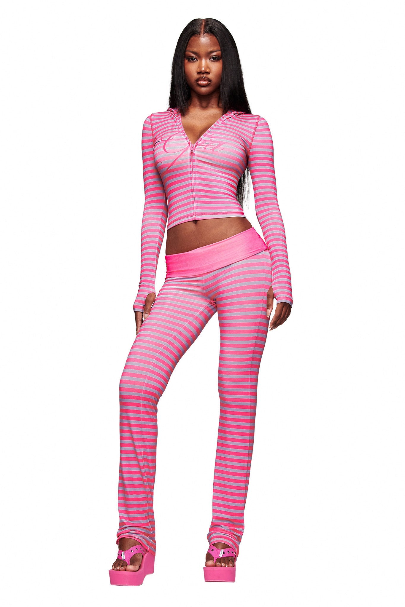 A woman stands confidently in I.AM.GIA’s BLARE FOLDOVER TRACKPANT in hot pink stripe, paired with a matching long-sleeve zip-up top and pink platform sandals, set against a plain white background.