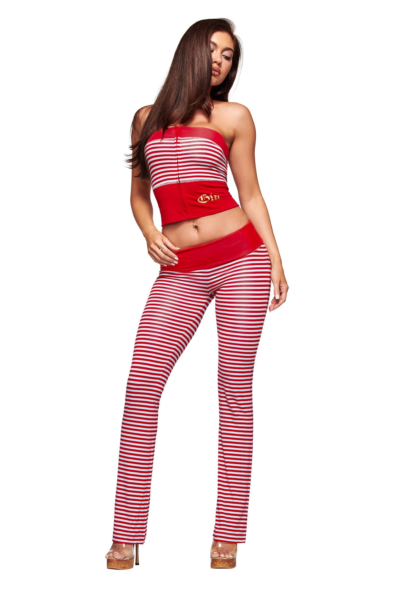 A woman poses confidently in an I.AM.GIA BLARE FOLDOVER TRACKPANT - RED AND BLUE STRIPE, made from soft viscose, paired with a red strapless crop top and tan heels against a white background.