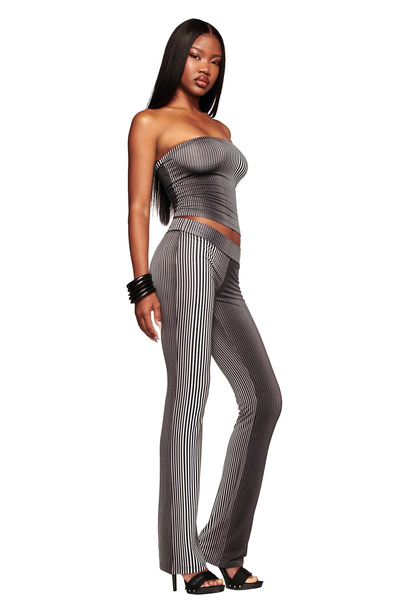 A woman poses confidently in I.AM.GIA's TRINITY 2-IN-1 SKIRT PANT - STRIPE, featuring vertical black and white stripes. She pairs it with a strapless fitted top, high heels, black bangles, and has long straight hair against a white background.