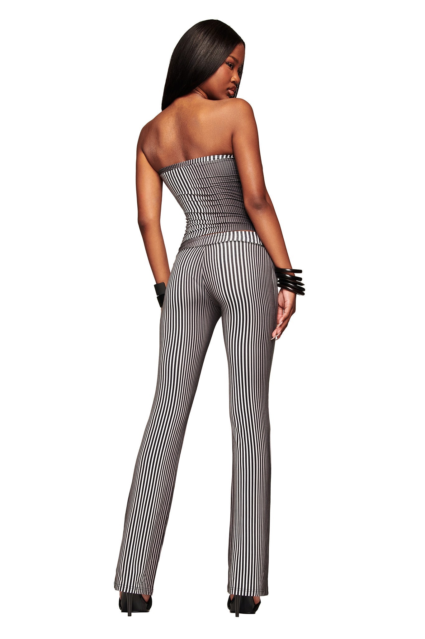 A woman with long hair down stands facing away, looking over her shoulder, wearing the I.AM.GIA TRINITY 2-IN-1 SKIRT PANT - STRIPE and black high heels.