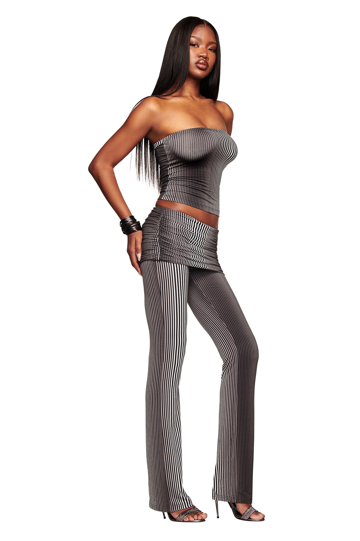 A woman poses in the I.AM.GIA TRINITY 2-IN-1 SKIRT PANT - STRIPE, a black and white ombre stripe set, styled with black bracelets and open-toe heels. She has long straight hair and stands against a white background.