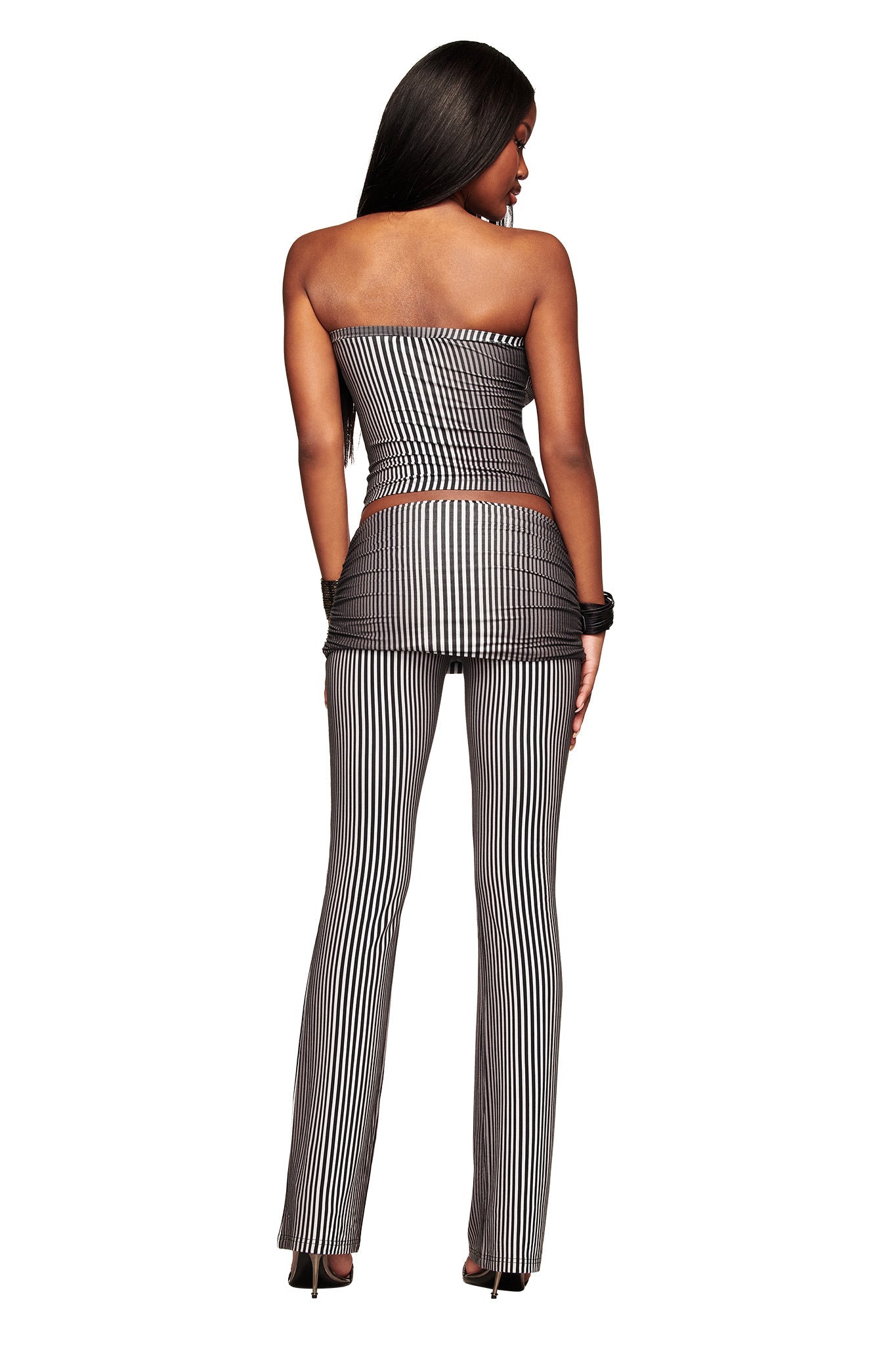 A woman with long dark hair stands facing away, wearing the I.AM.GIA TRINITY BOOB TUBE - STRIPE top and matching flared pants in black and white, styled with black heels and a bracelet against a white background.