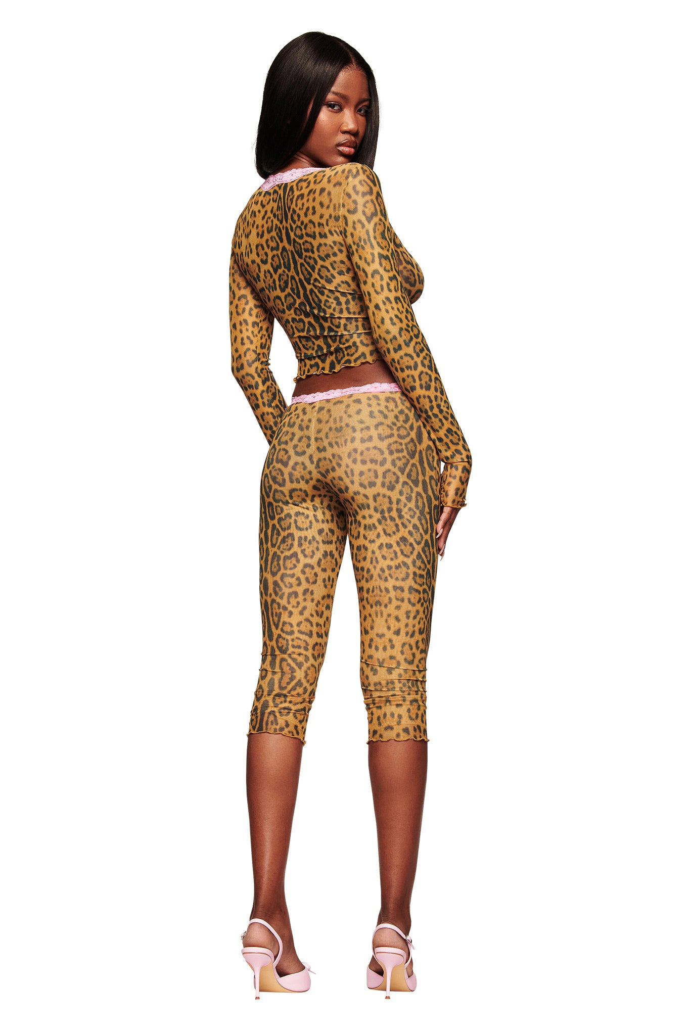 A woman faces away, turning her head slightly toward the camera. She wears the I.AM.GIA TASHI TOP - PANTHERA with matching knee-length leggings featuring pink trim and white high-heeled sandals, set against a white background.