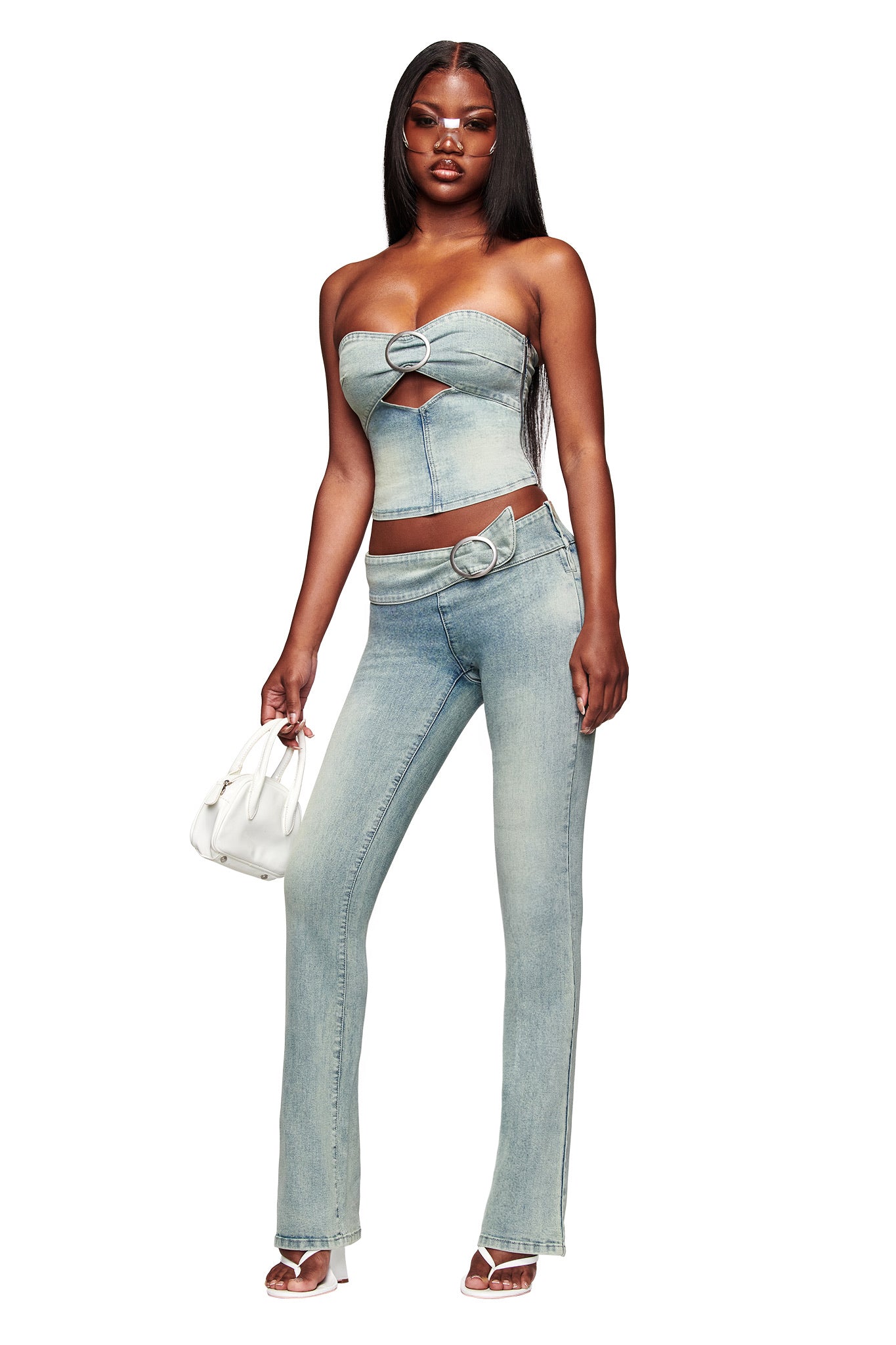 A woman holds a small white purse and wears the I.AM.GIA JENN DENIM BOOB TUBE - BLUE, paired with matching high-waisted jeans featuring a ring belt, plus white heeled sandals.