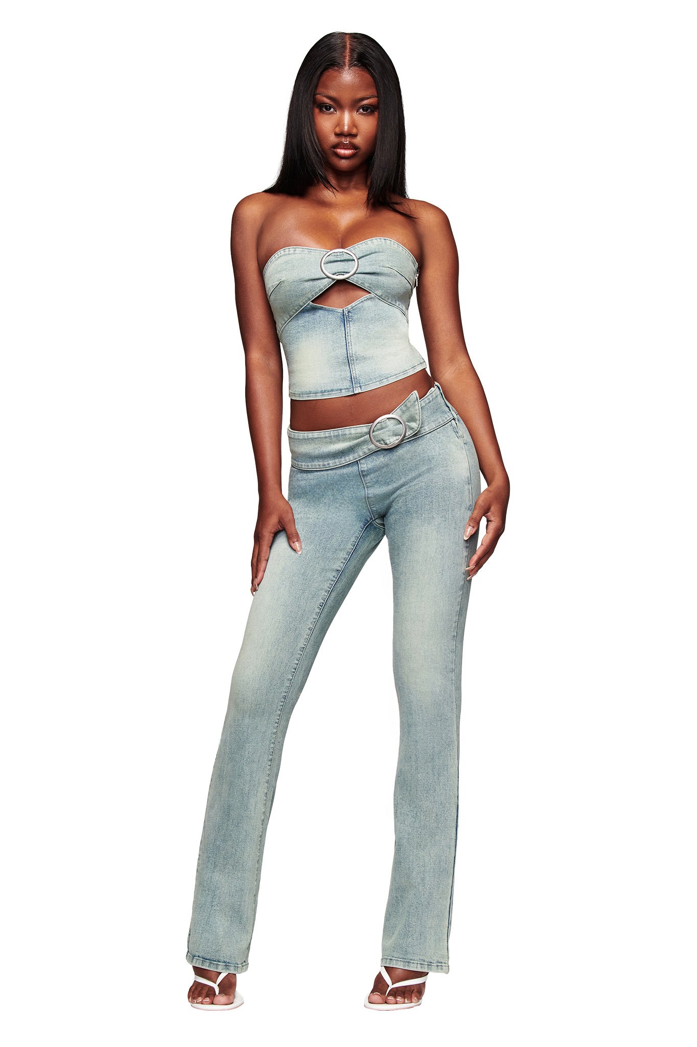 A woman stands confidently in the I.AM.GIA JENN JEAN - BLUE set: a strapless light blue denim crop top with a heart-shaped cutout and matching mid-rise flared pants, styled with clear-heeled sandals.