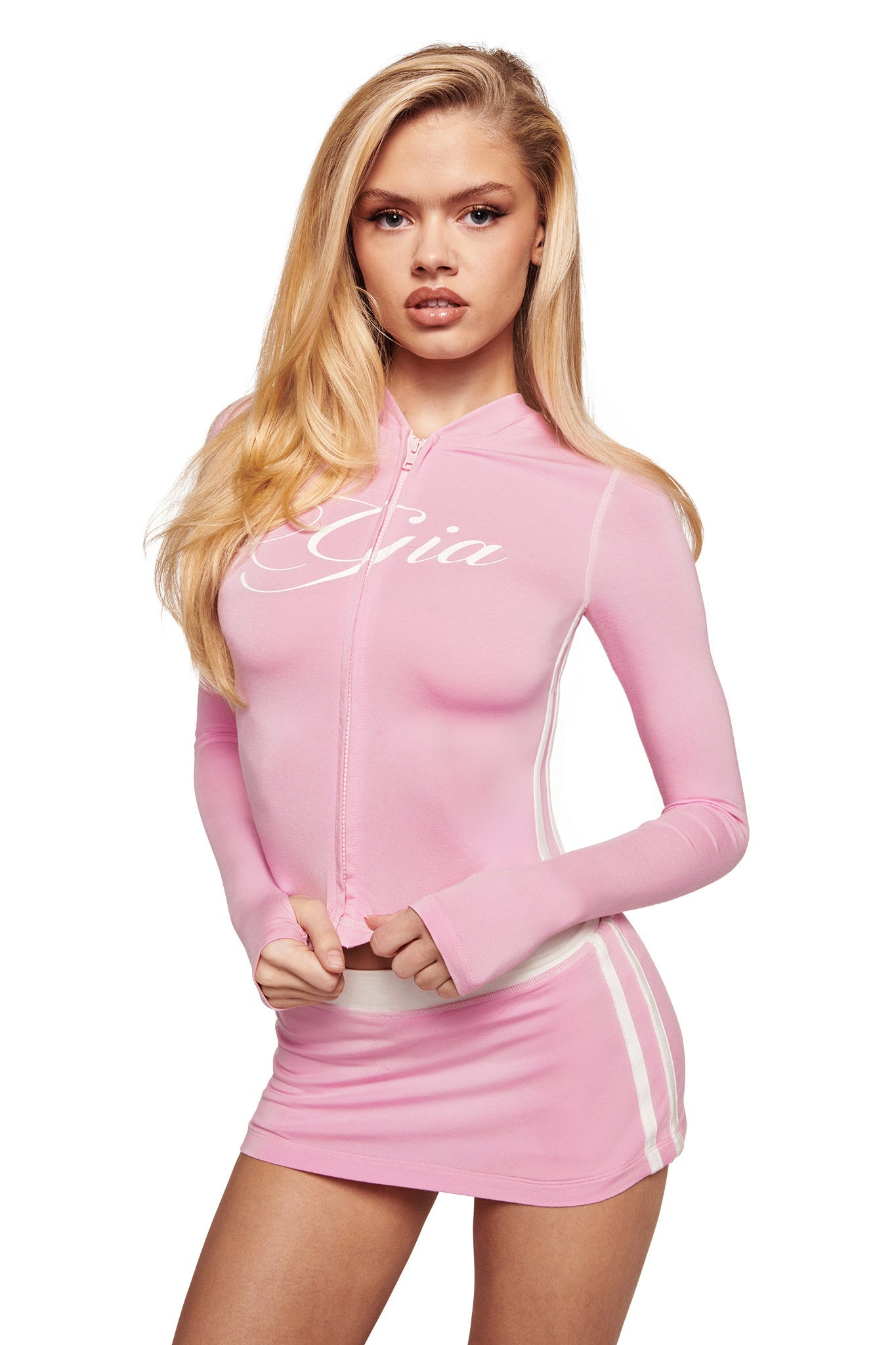 A woman with long blonde hair wears the I.AM.GIA BLARE PIPING HOODIE in baby pink and white, paired with a matching mini skirt. She stands against a white background, holding the bottom of her hoodie.