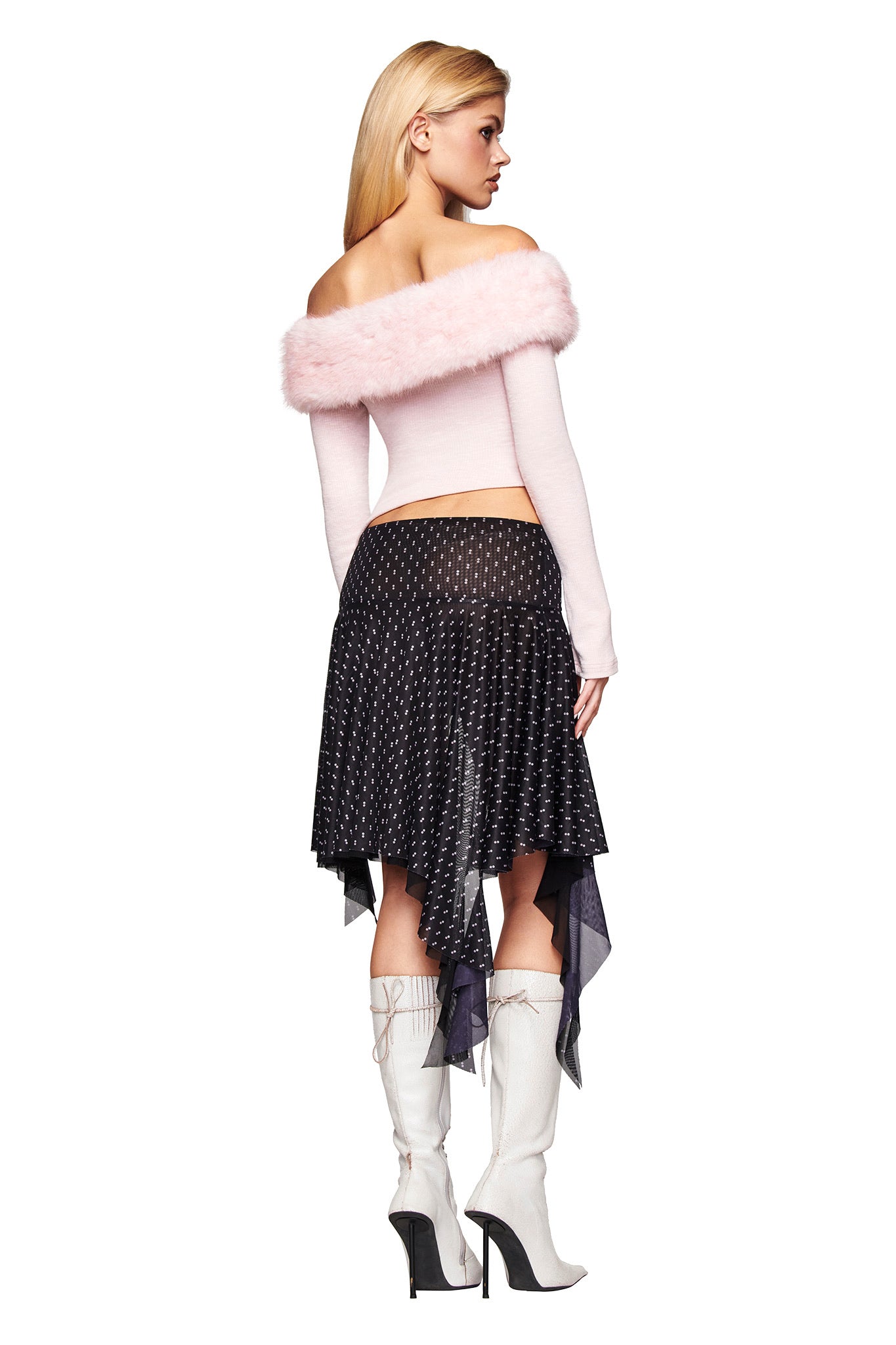 A woman stands facing away, wearing the I.AM.GIA CECILY TOP in pink with a faux fur collar, a black asymmetrical dotted skirt, and white knee-high heeled boots. Her long blonde hair is down.