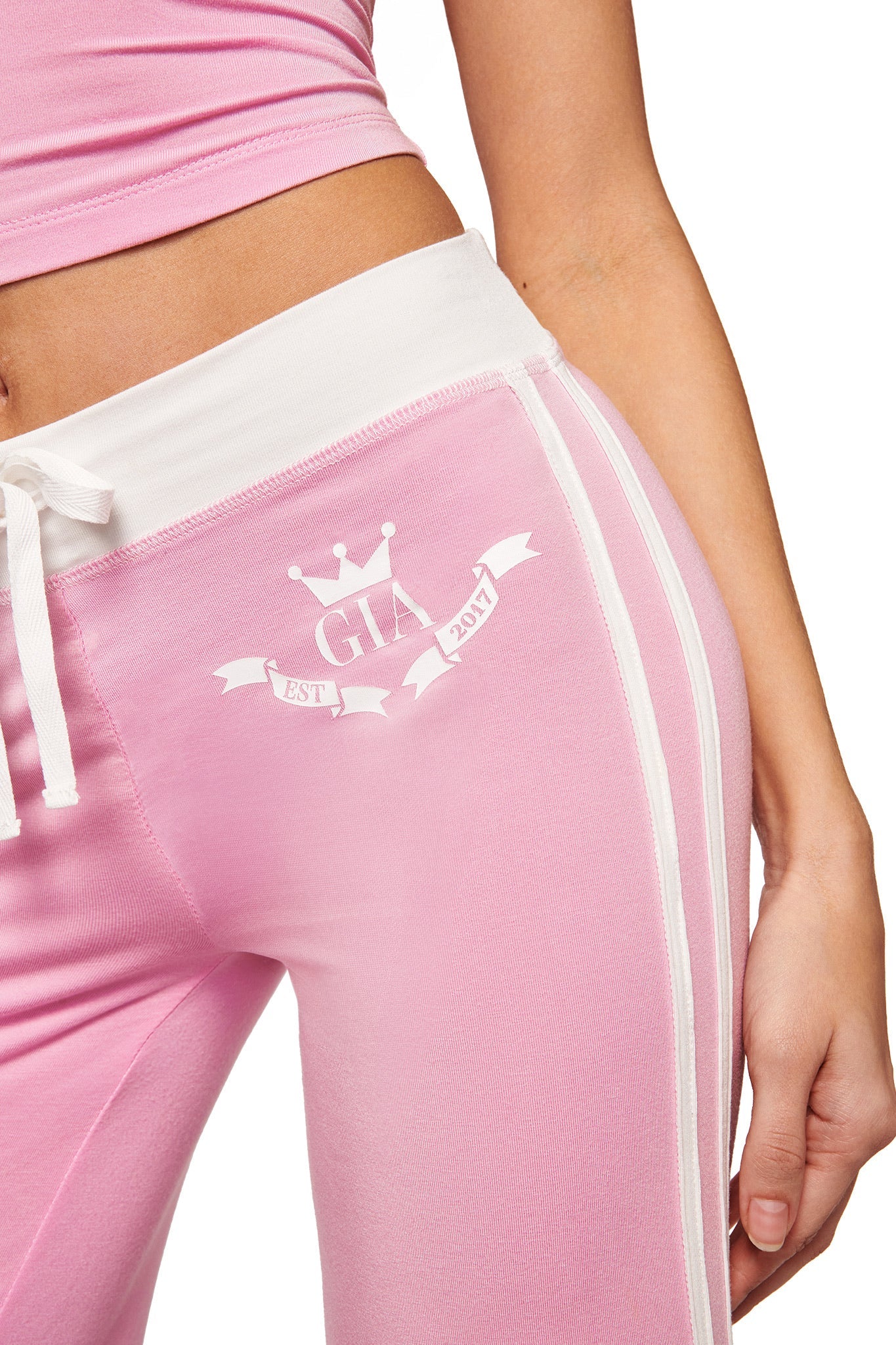 A person models the I.AM.GIA BLARE PIPING TRACKPANT in baby pink with white stripes and waistband. "GIA" and "EST. 2017" with a crown and banner appear on the hip. Only the midsection and part of one arm are shown.