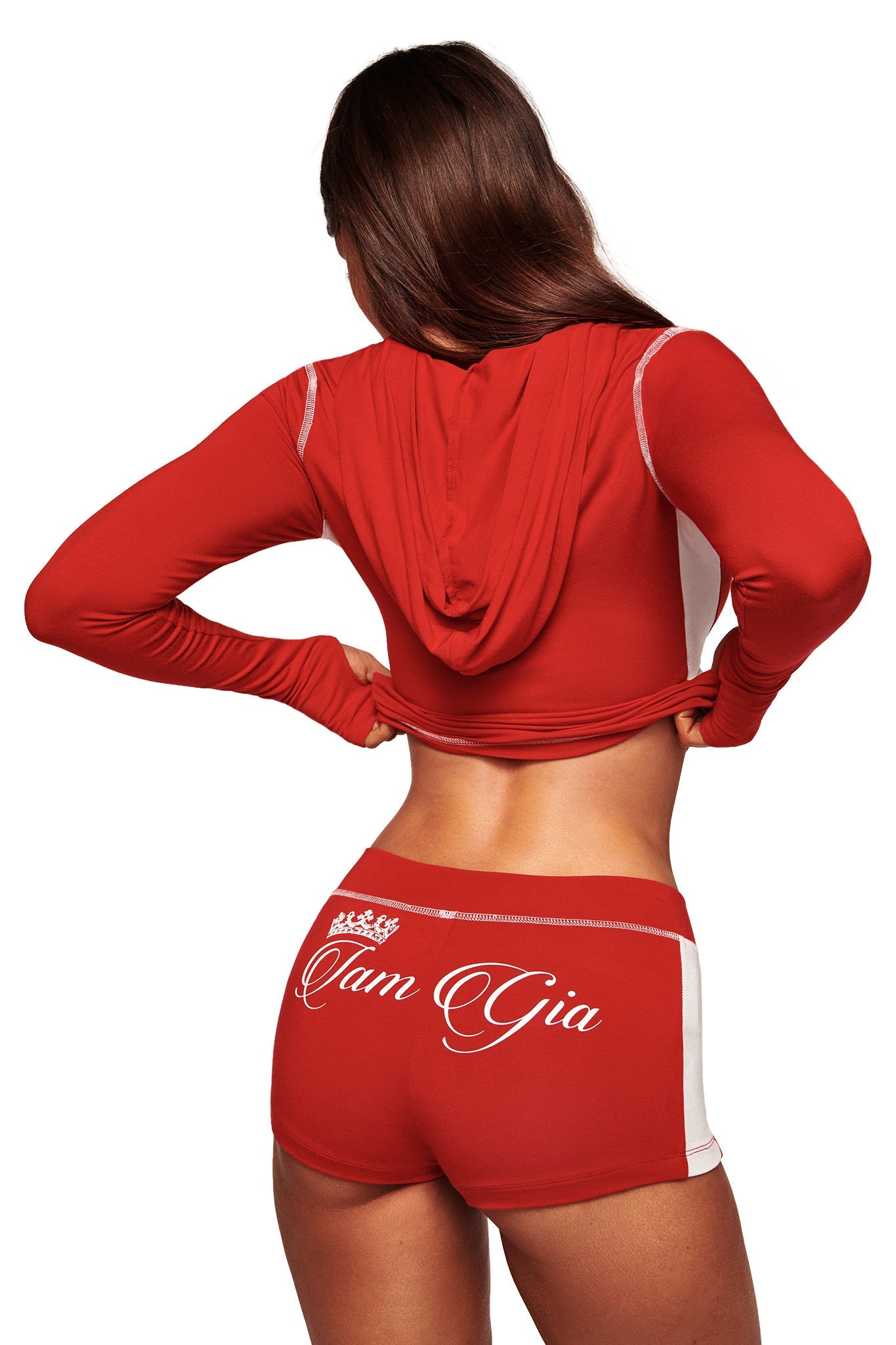 A woman with long brown hair is seen from behind, slightly lifting her BLARE PANELLED HOODIE in red by I.AM.GIA, paired with matching sporty shorts featuring white trim and "I am Gia" on the back.