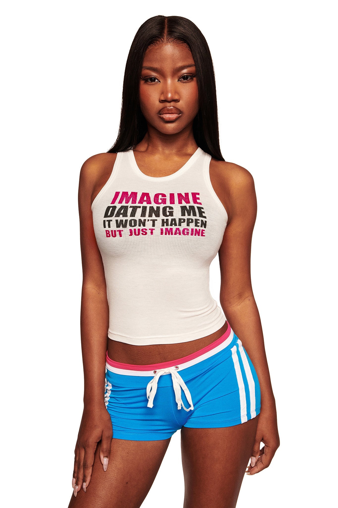 A woman with long straight hair wears the I.AM.GIA POLINA TANK - IMAGINE, a white racer-style rib knit tank top with "IMAGINE DATING ME IT WON'T HAPPEN BUT JUST IMAGINE" text, paired with blue athletic shorts. She stands facing forward.