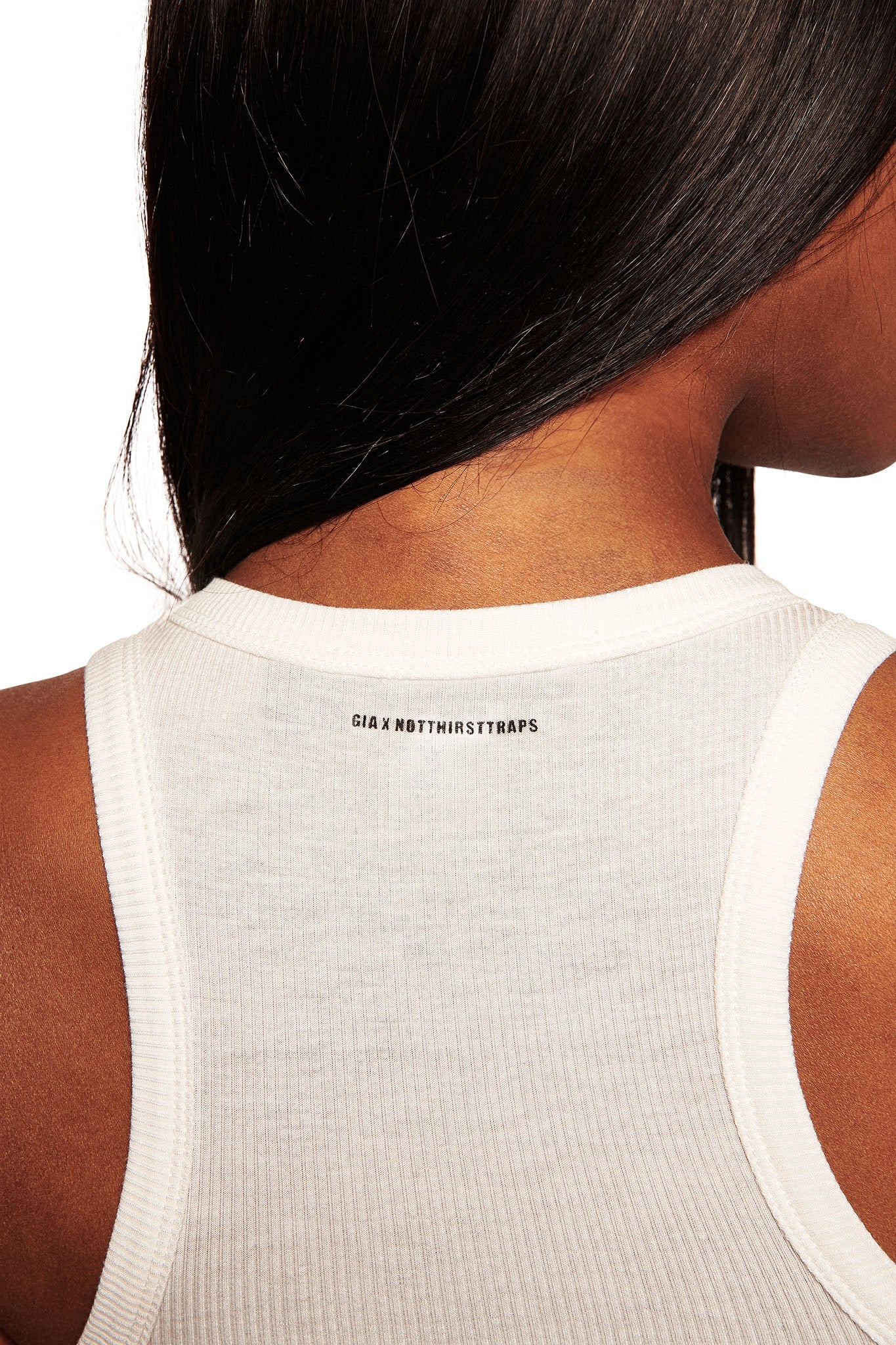 Close-up of a person wearing the I.AM.GIA POLINA TANK - MANIPULATOR, a rib knit tank with “GIA X N0T4HRSTRAP5” printed in black below the neckline. Their long black hair sweeps to one side, highlighting the racerback design.