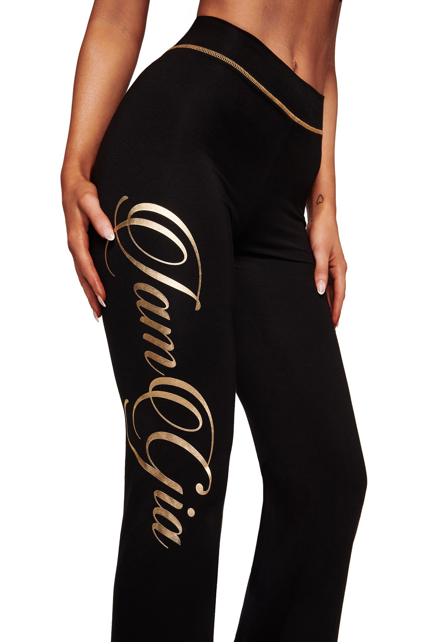 A person wears the I.AM.GIA BLARE FORM TRACKPANT in black and gold, featuring a large metallic gold "I.AM.GIA" print down the left leg, with their hand resting on their hip.