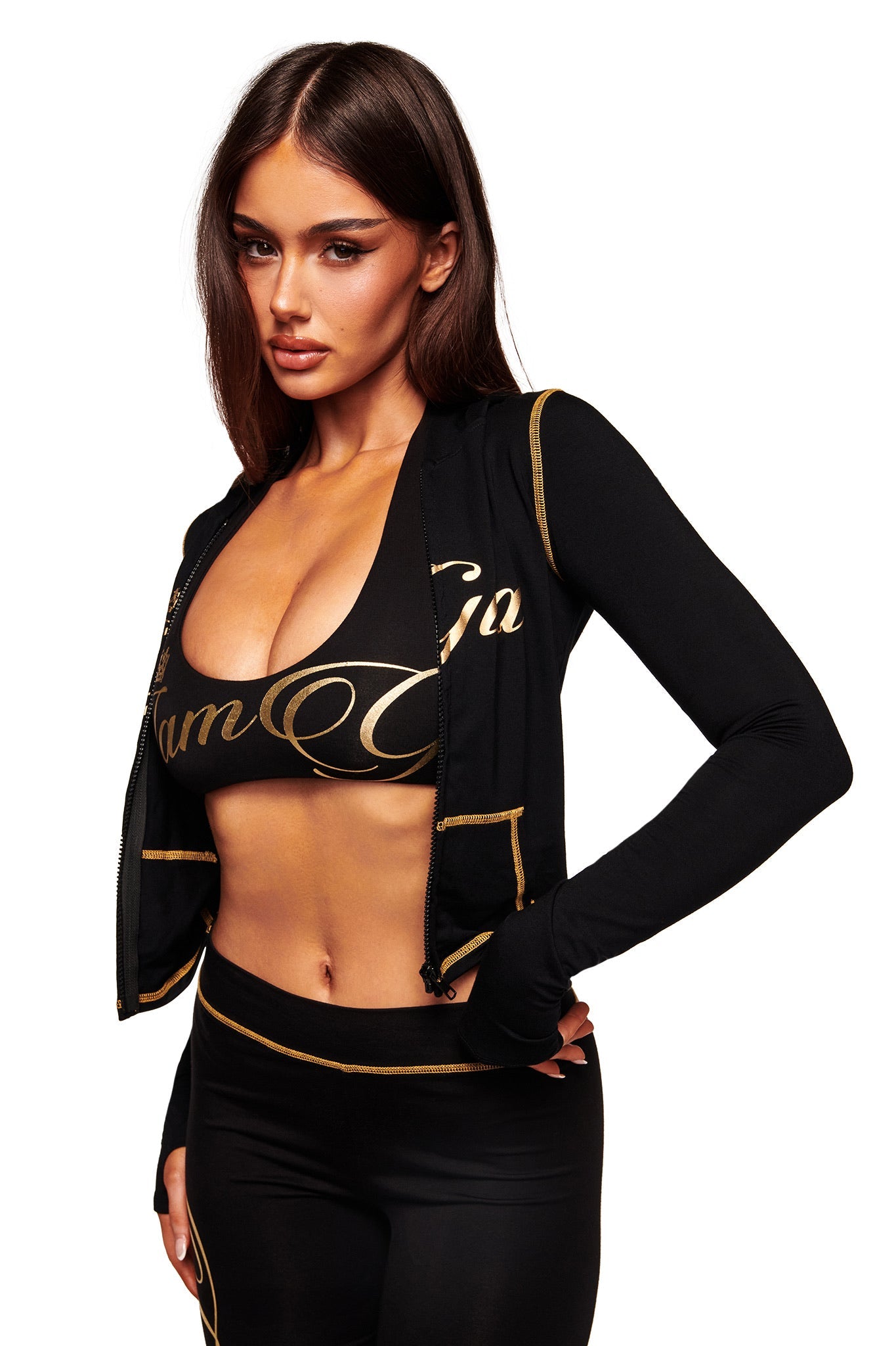 A woman with long brown hair models the I.AM.GIA BLARE FORM MULTI POCKET HOODIE in black and gold, unzipped to show bold gold lettering, over matching sportswear as she poses against a plain white background.