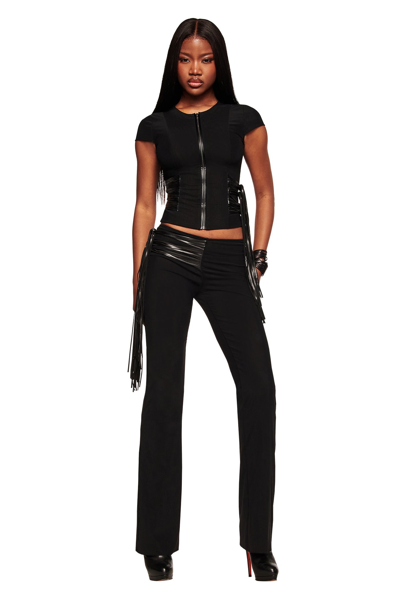 A woman faces forward in the I.AM.GIA ISELDA FRINGE TOP - BLACK and black pants with faux leather fringe details. She has long, straight hair, wears black platform heels, and stands against a plain white background.