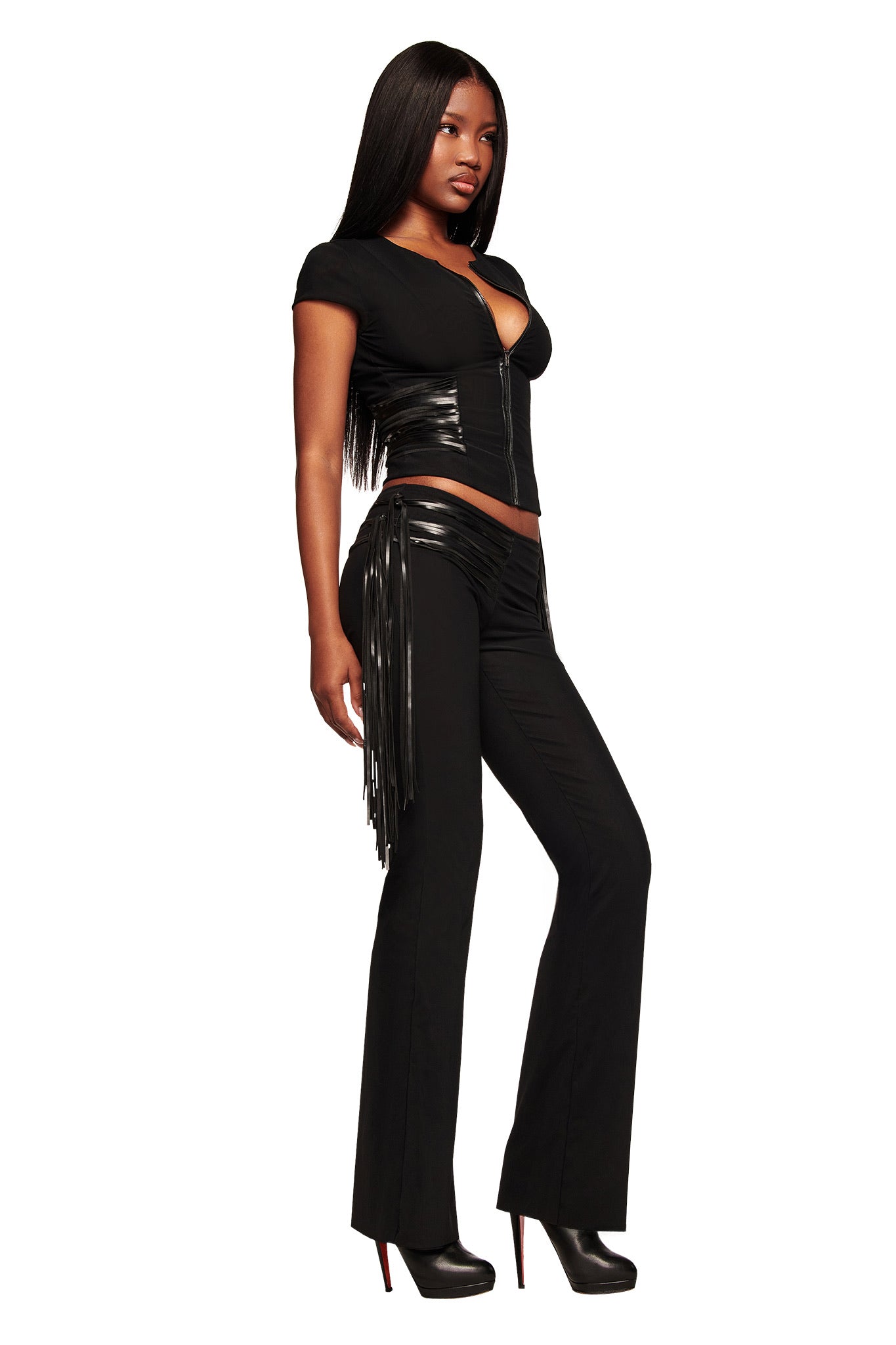 A woman with long straight hair models the I.AM.GIA ISELDA FRINGE TOP in black, featuring shiny faux leather fringe at the hips. She pairs it with black high heels and poses confidently against a white background.