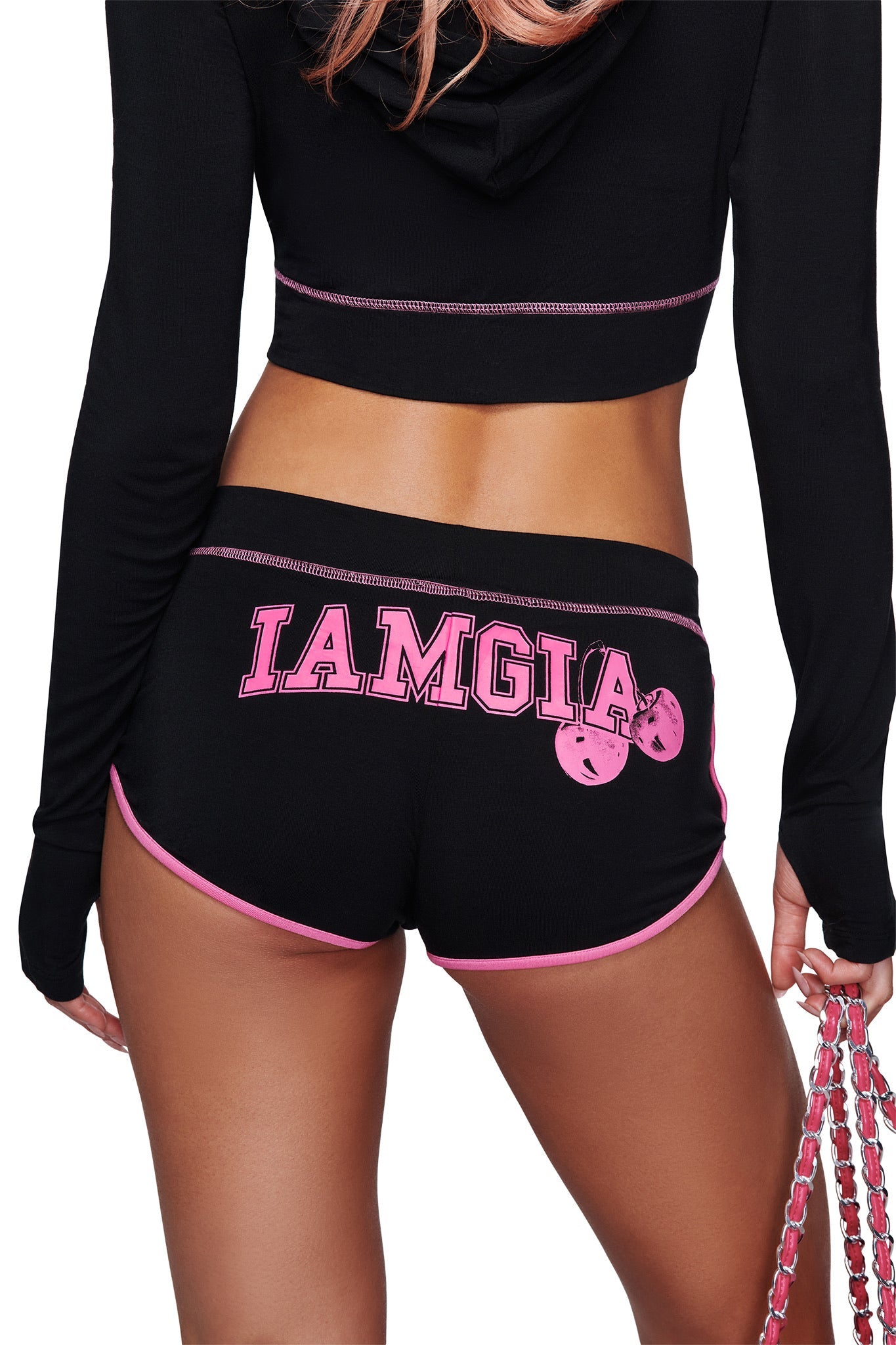 A woman wearing the BLARE DOLPHIN SHORT in black by I.AM.GIA, featuring pink trim and "I.AM.GIA" printed on the back, holds a pink chain accessory in her right hand.