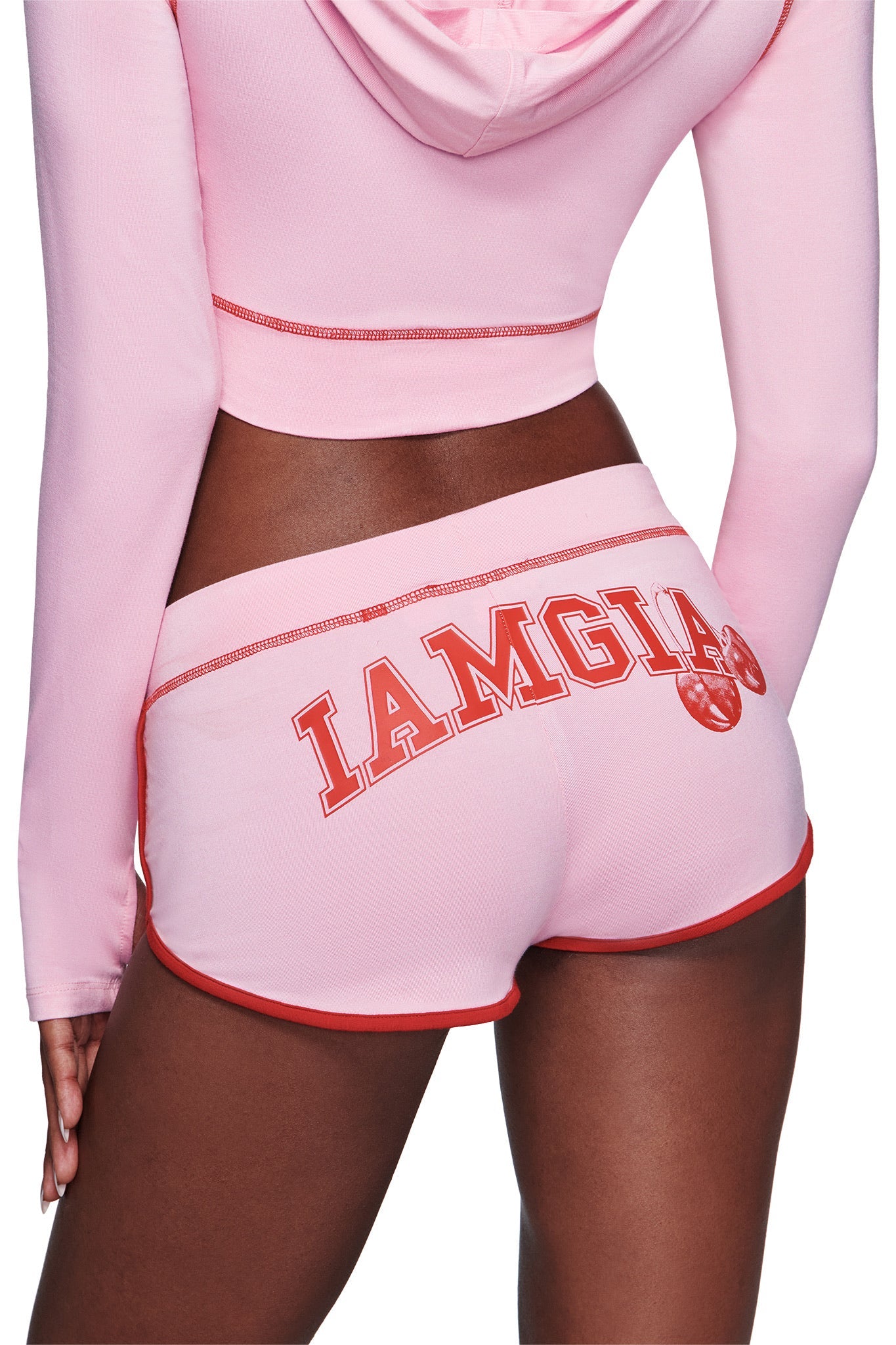 Dressed in the BLARE DOLPHIN SHORT - BABY PINK by I.AM.GIA, featuring bold red trim and "IAMGIA" lettering on the back, this trendy bamboo dolphin short pairs flawlessly with a pink cropped hoodie for an effortlessly cool summer beachwear vibe.
