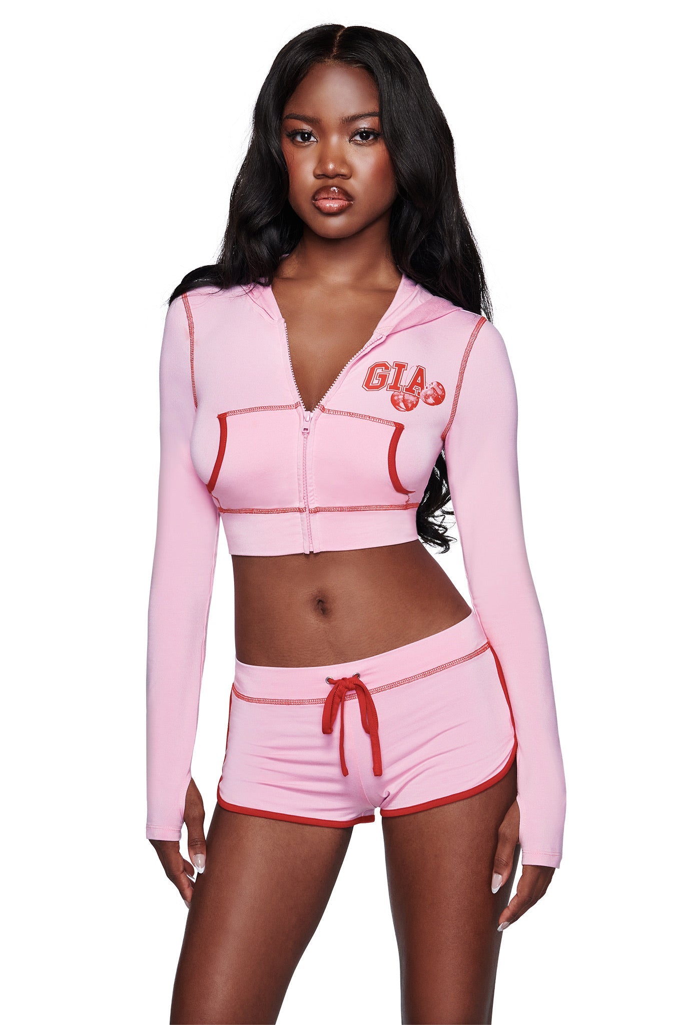 A woman stands facing forward wearing the BLARE DOLPHIN SHORT in baby pink from I.AM.GIA, paired with a light pink crop hoodie featuring red trim and "GIA" printed on the left chest. Her long dark hair and neutral expression highlight this stylish set, ideal for summer beachwear.