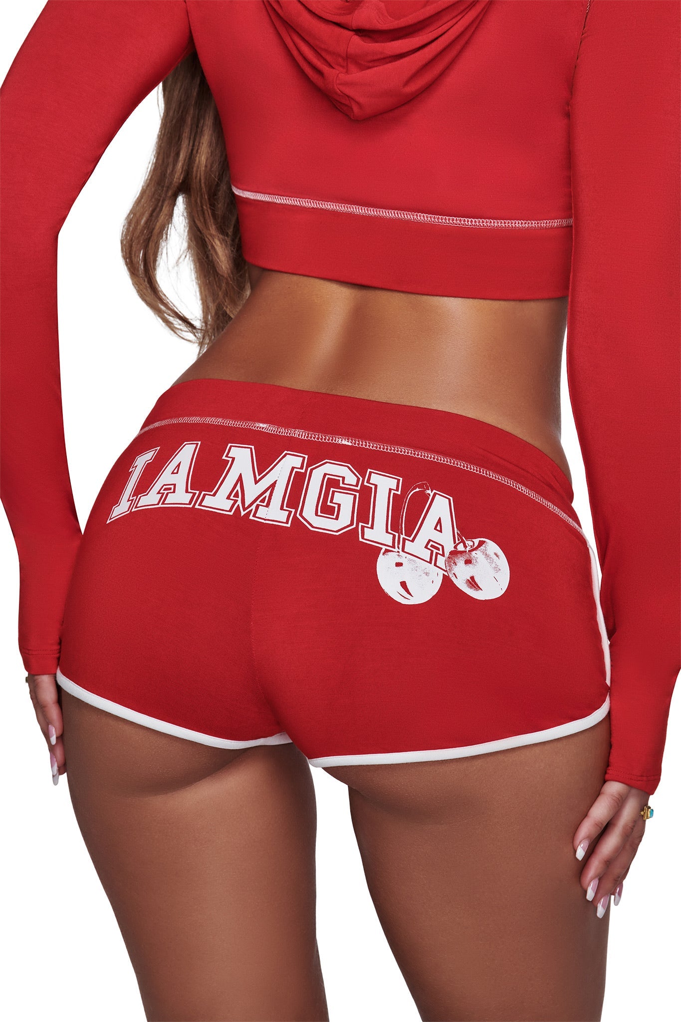 A woman wearing a red crop hoodie and the I.AM.GIA BLARE DOLPHIN SHORT in red, featuring white trim. The shorts have “LAMGIA” in white text and a graphic of two white cherries on the back, reminiscent of the brand’s iconic style.