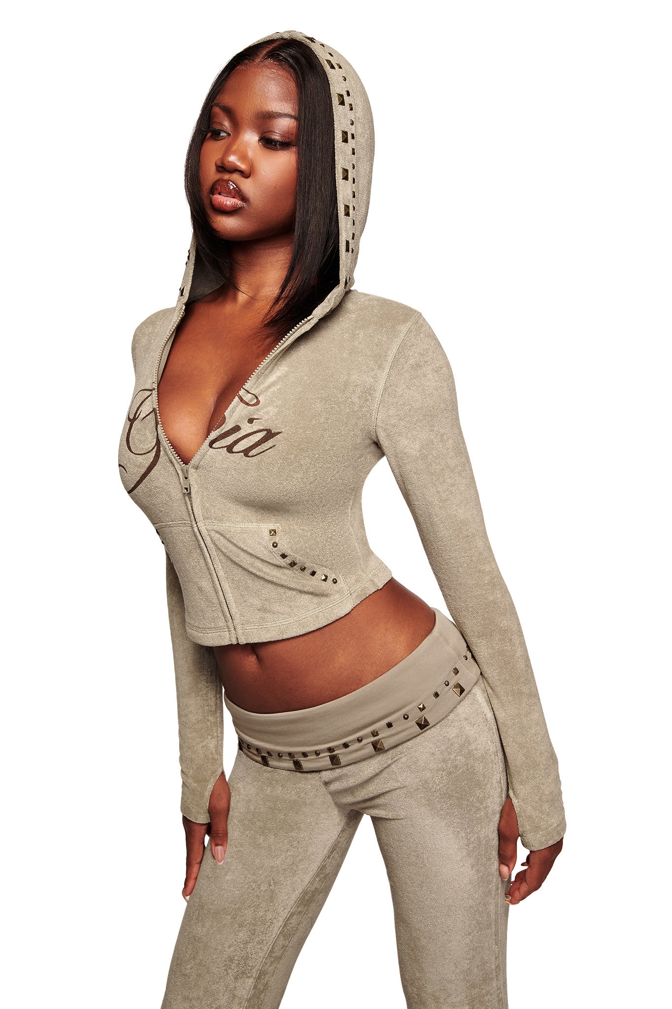 A woman poses against a white background in the I.AM.GIA BLARE STUDDED TERRY HOODIE in Stone, a beige velour zip-up with studded details and script lettering, paired with matching low-rise pants.