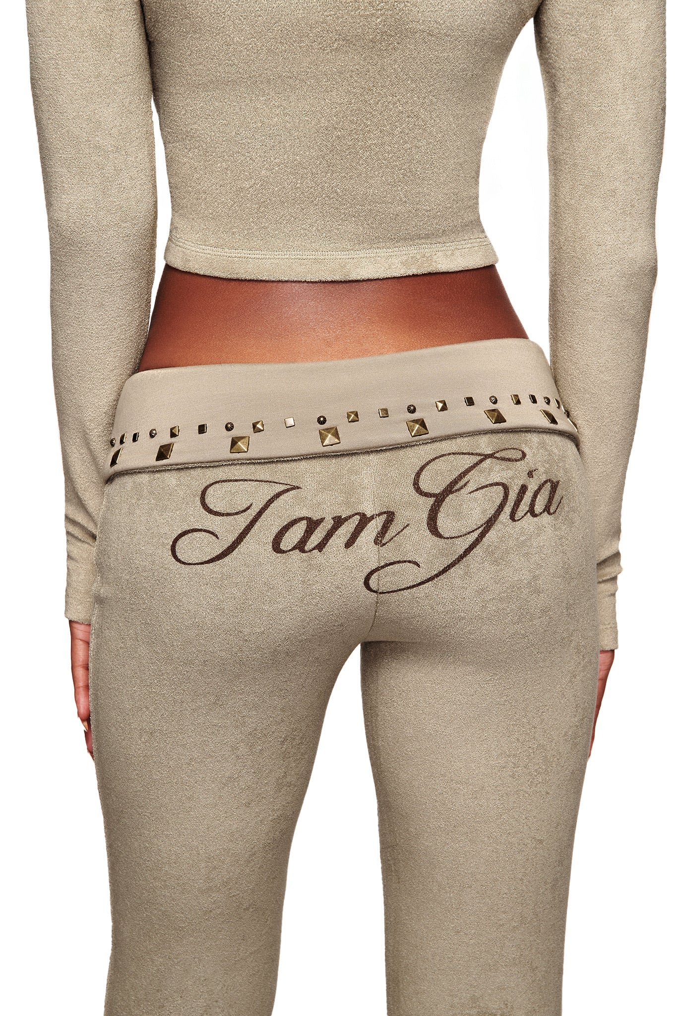 A person seen from the back wears matching beige I.AM.GIA BLARE STUDDED TERRY PANTS, featuring "I am Gia" in bold script and a studded metallic waistband.
