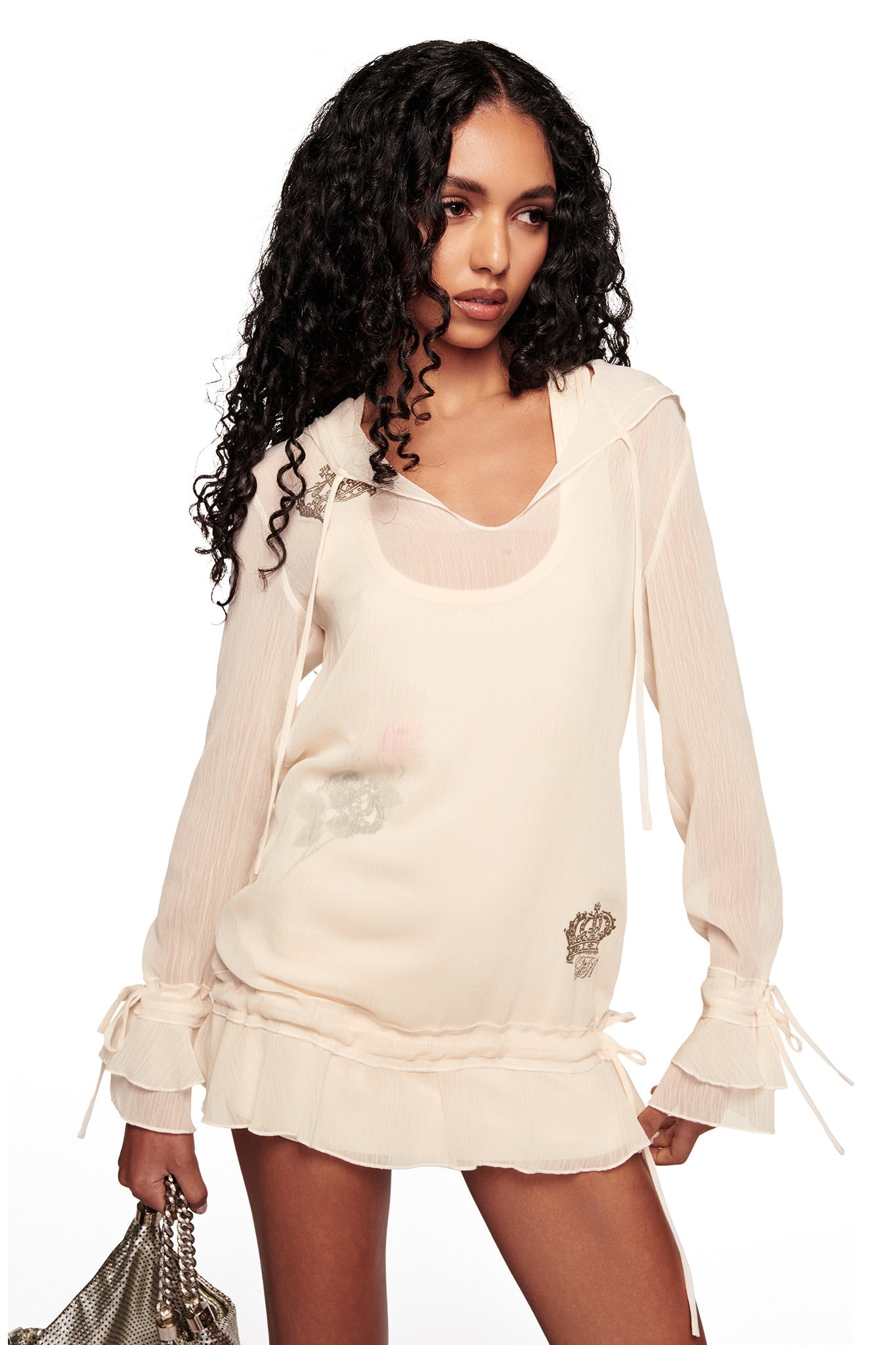 A woman with long curly hair wears the I.AM.GIA DIAMOND 2-IN-1 MINI DRESS in cream, featuring ruffle details at the hem and cuffs, and holds a metallic bag while posing against a white background.