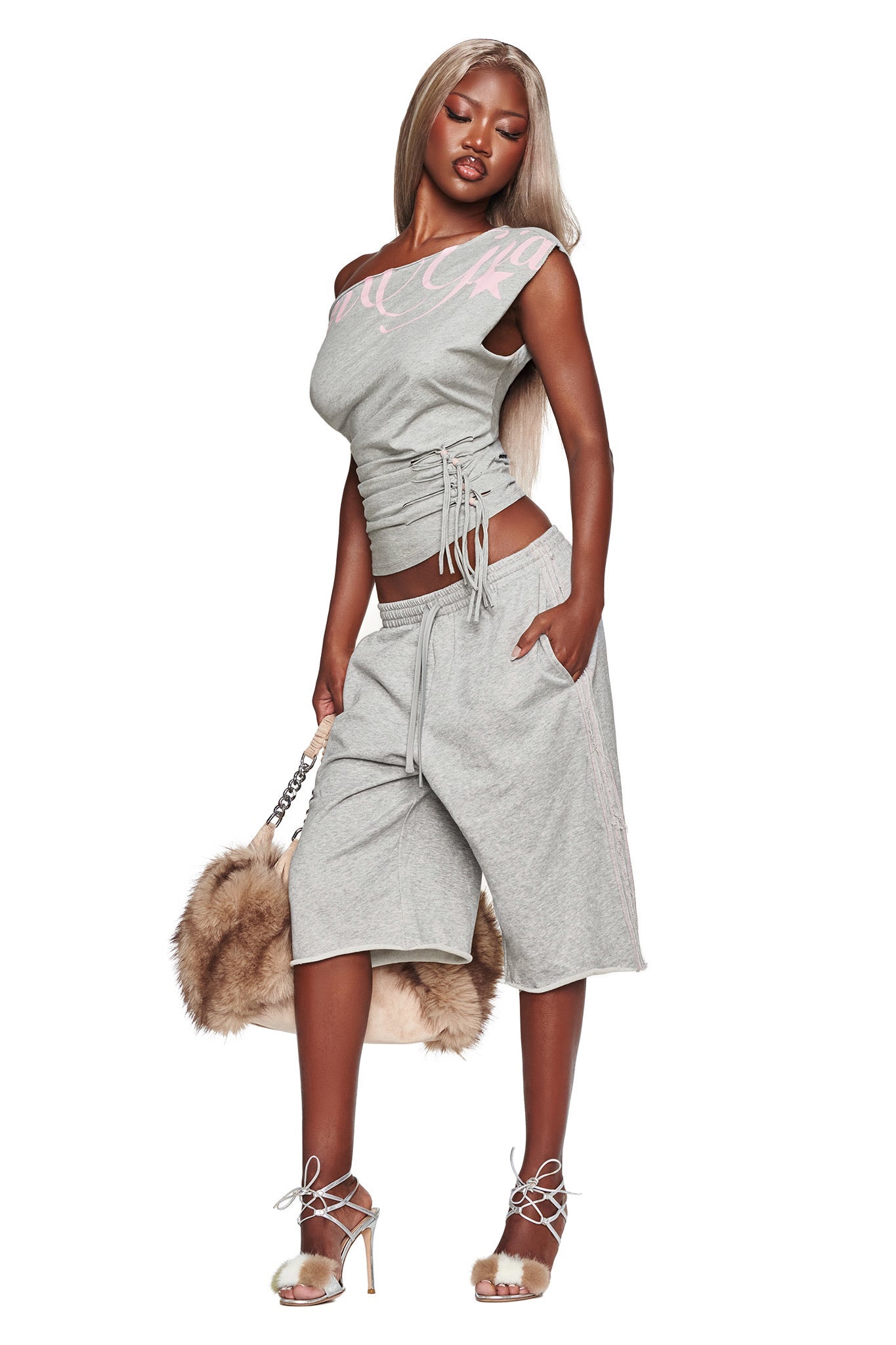 Wearing the BEX OFF SHOULDER TOP in grey by I.AM.GIA, this woman poses against a white background. The gray off-shoulder top features a ruched bodice with side ruching and is paired with matching long shorts. She accessorizes with a brown faux fur bag and strappy heels.