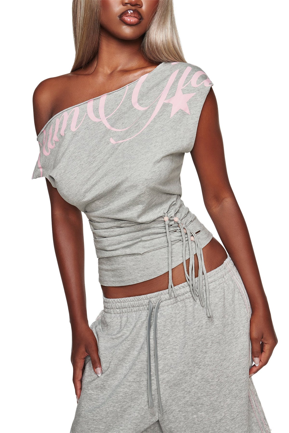 A woman models the I.AM.GIA BEX OFF SHOULDER TOP in grey, featuring pink lettering, paired with matching grey drawstring pants as she poses with one hand on her hip against a white background.