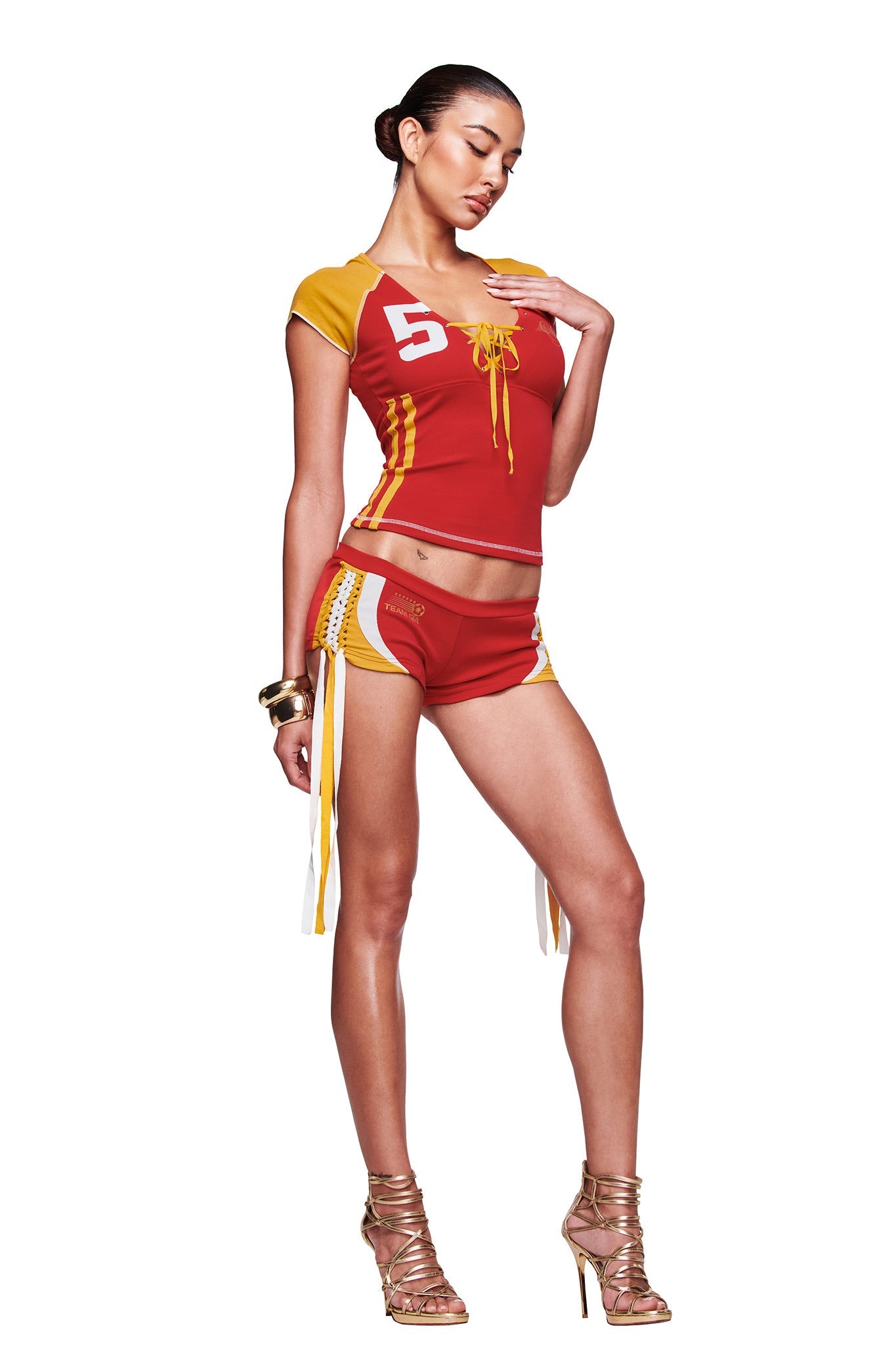 A woman poses in the I.AM.GIA NOVIA MICRO SPORT SHORT - RED, paired with a matching sporty top featuring the number 5 and lace-up details. She wears high heels, has her hair pulled back, and stands against a white background.