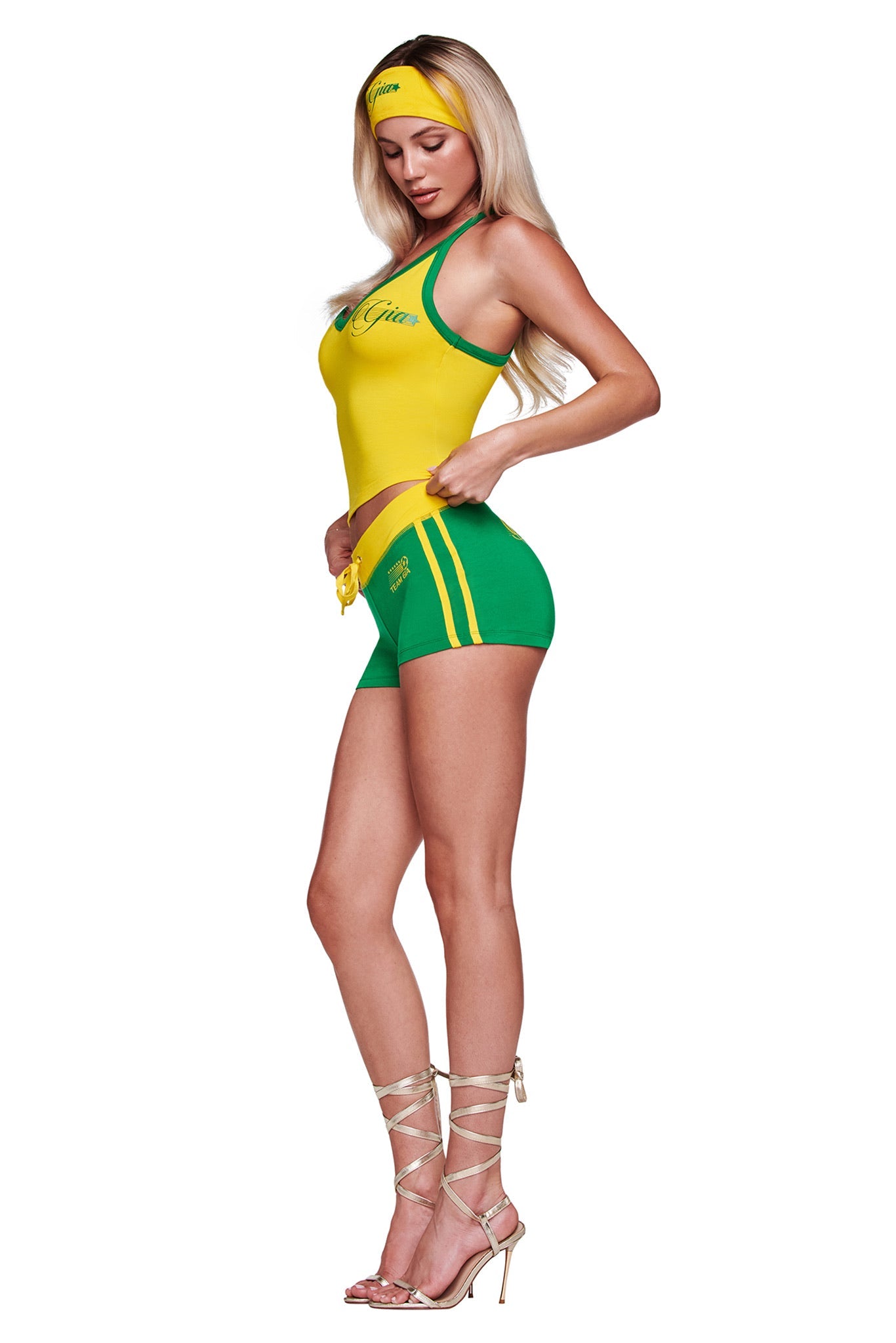 A woman with long blonde hair stands in profile against a white background, looking down as she adjusts her I.AM.GIA BLARE PIPING SHORT in green, paired with a yellow and green sporty outfit, matching headband, and strappy high heels.