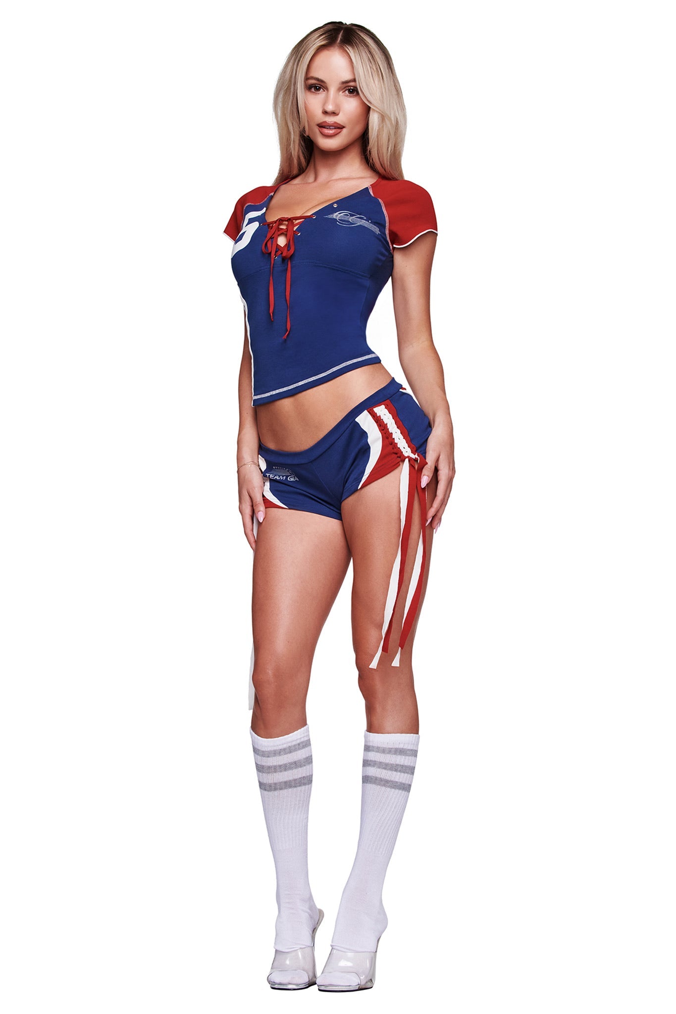 A woman poses against a white background in I.AM.GIA's NOVIA MICRO SPORT SHORT - NAVY, styled with a cheerleader-inspired blue and red crop top, white knee-high socks, and high heels.
