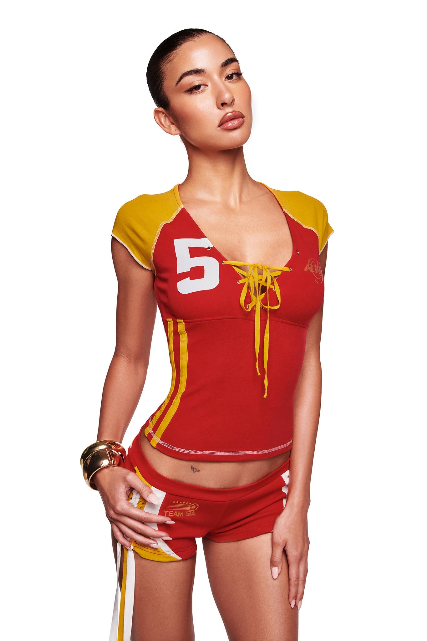 A woman poses against a white background wearing the I.AM.GIA NOVIA Lace-Up T-Shirt and Headband in red, styled with matching shorts and gold bangles.